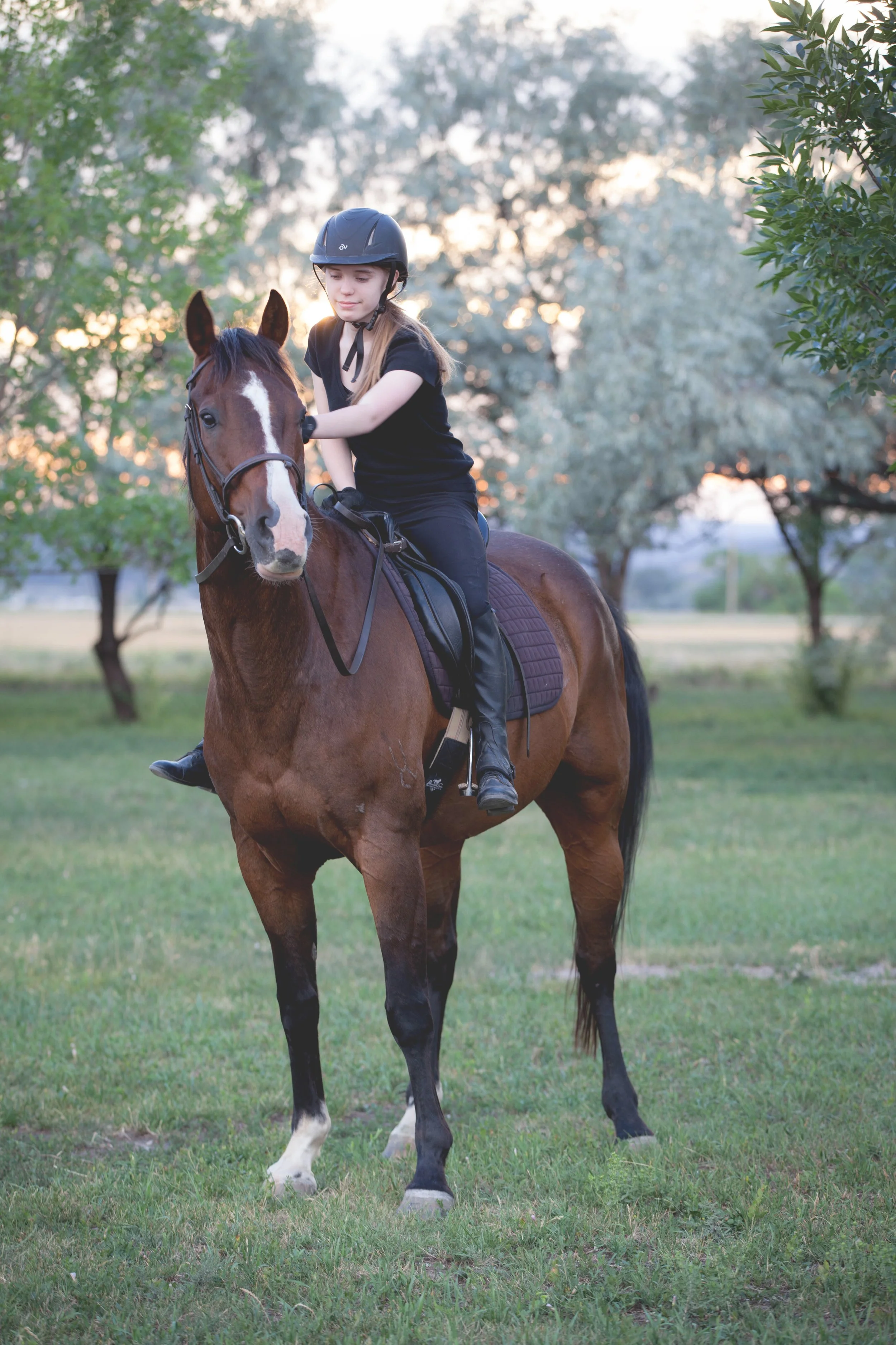 A young girl riding a brown horse in a grassy area with trees in the background during sunset.