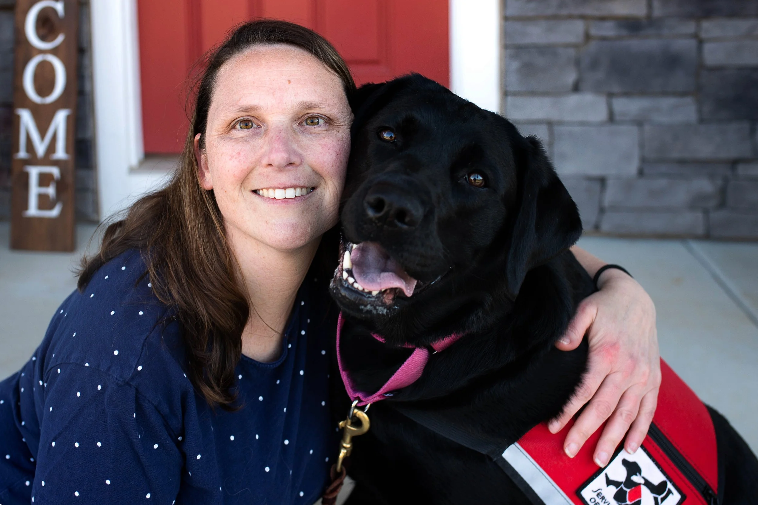 A woman with long brown hair and a blue polka dot shirt smiling and hugging a black service dog with a pink collar and red vest with a black and white logo, sitting in front of a house with a red door and a sign that says 'HOME' in the background.