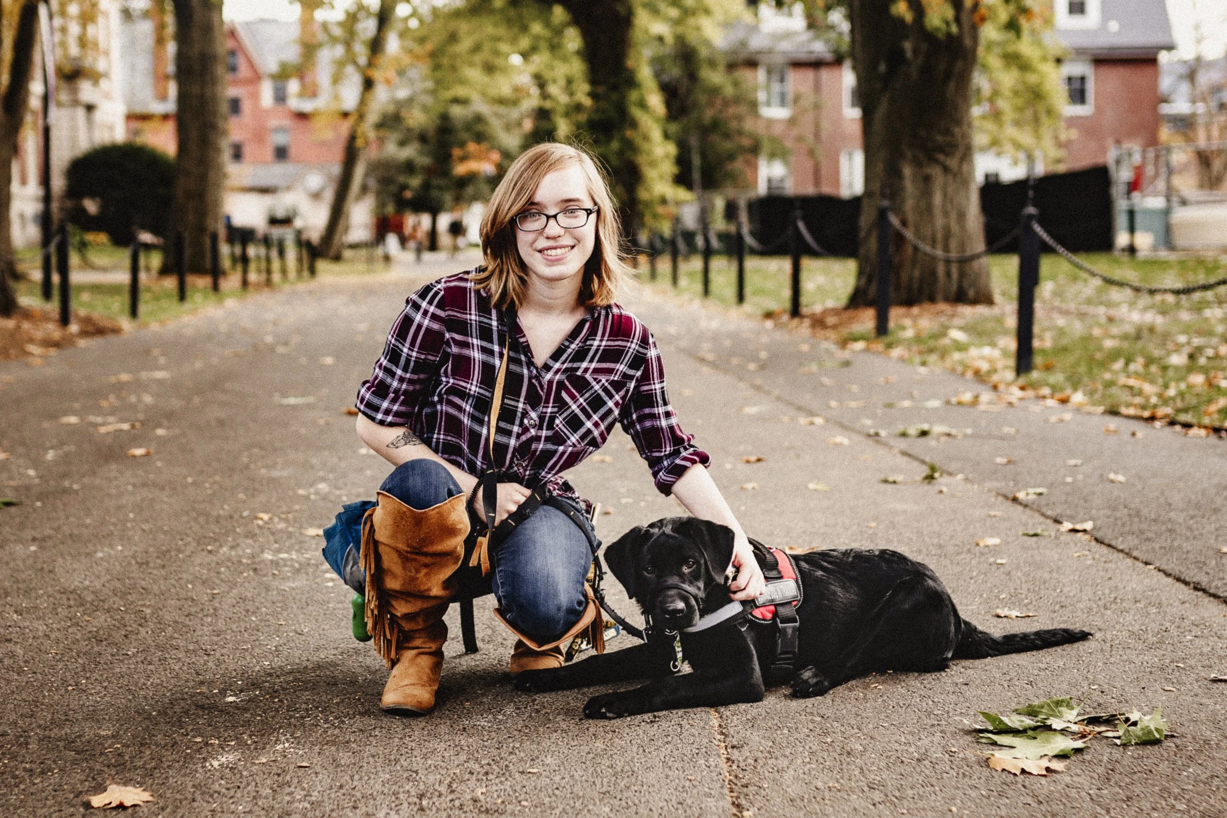 A woman with glasses, blonde hair, and a plaid shirt kneels on a park path, smiling, next to a black dog lying on the ground. The woman has a tattoo on her arm and is wearing brown boots. The park has trees, fallen leaves, and a chain-link fence in t