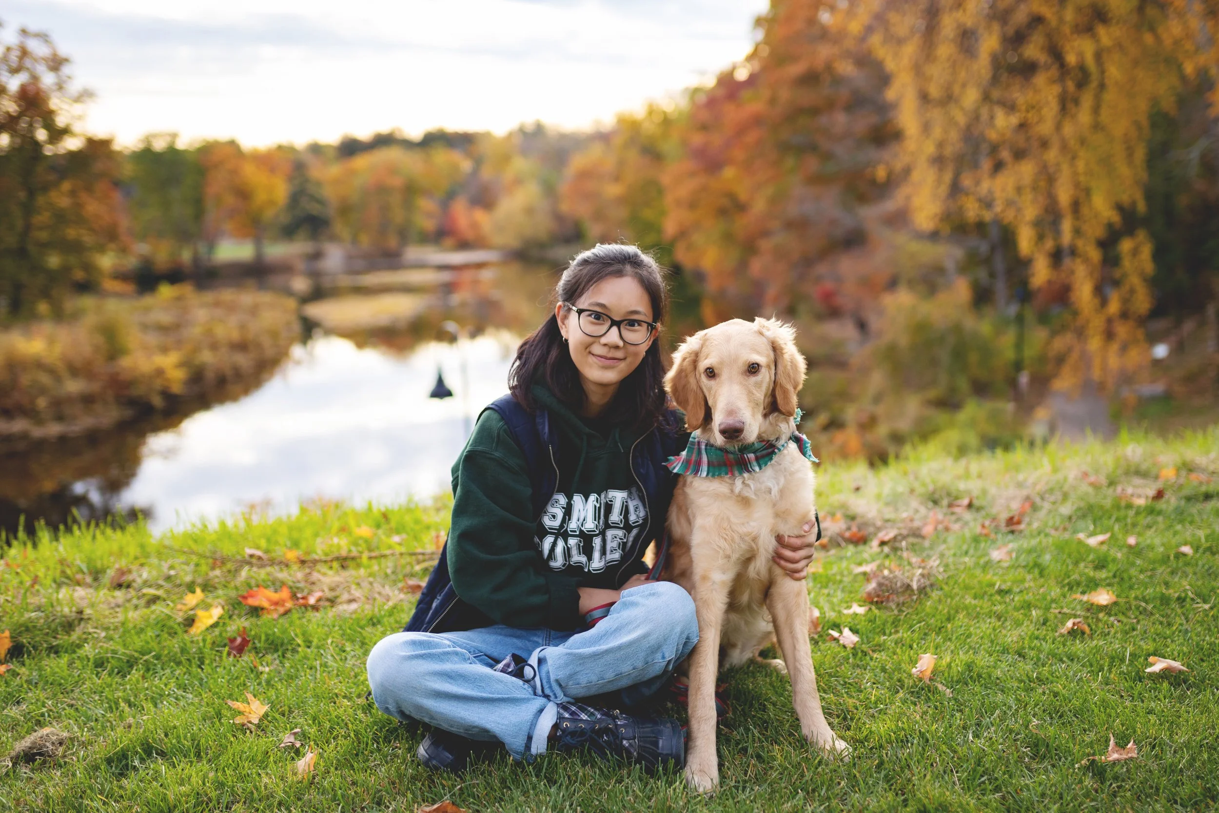 A young woman with glasses kneels on grass next to a golden retriever puppy near a lake with autumn trees in the background.