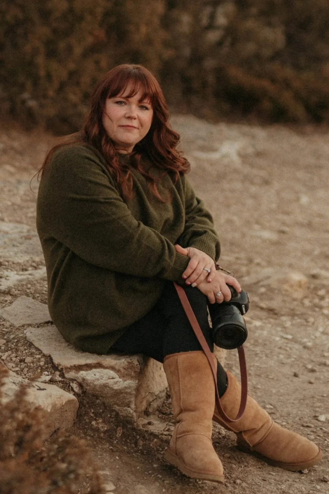 A woman with red hair, sitting on a rock on the beach, holding a camera, wearing a green sweater, black pants, and tan boots.