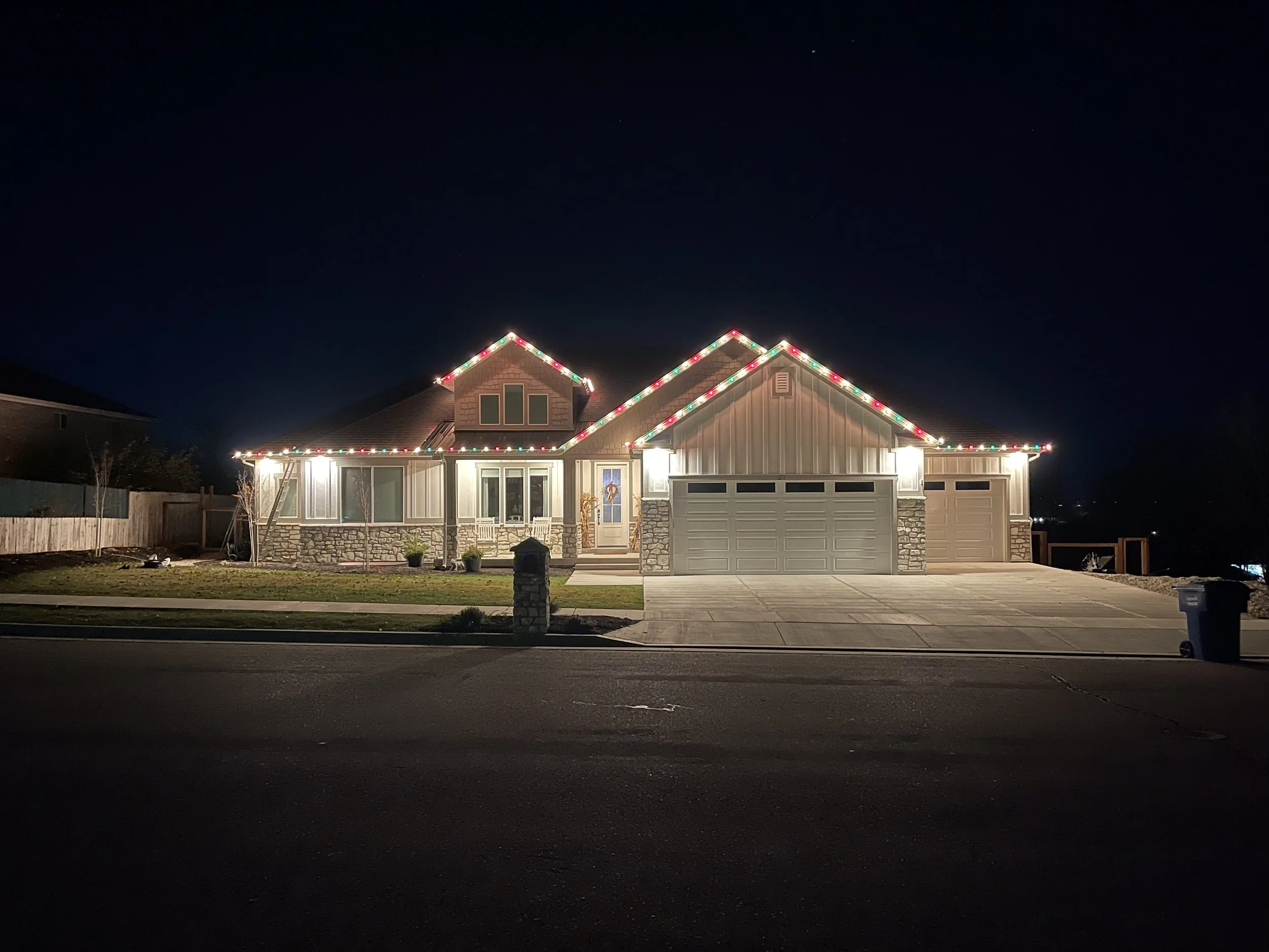 A house decorated with colorful Christmas string lights along the roofline at night. The house has stone and siding exterior, a three-car garage, and a small front porch with stairs.