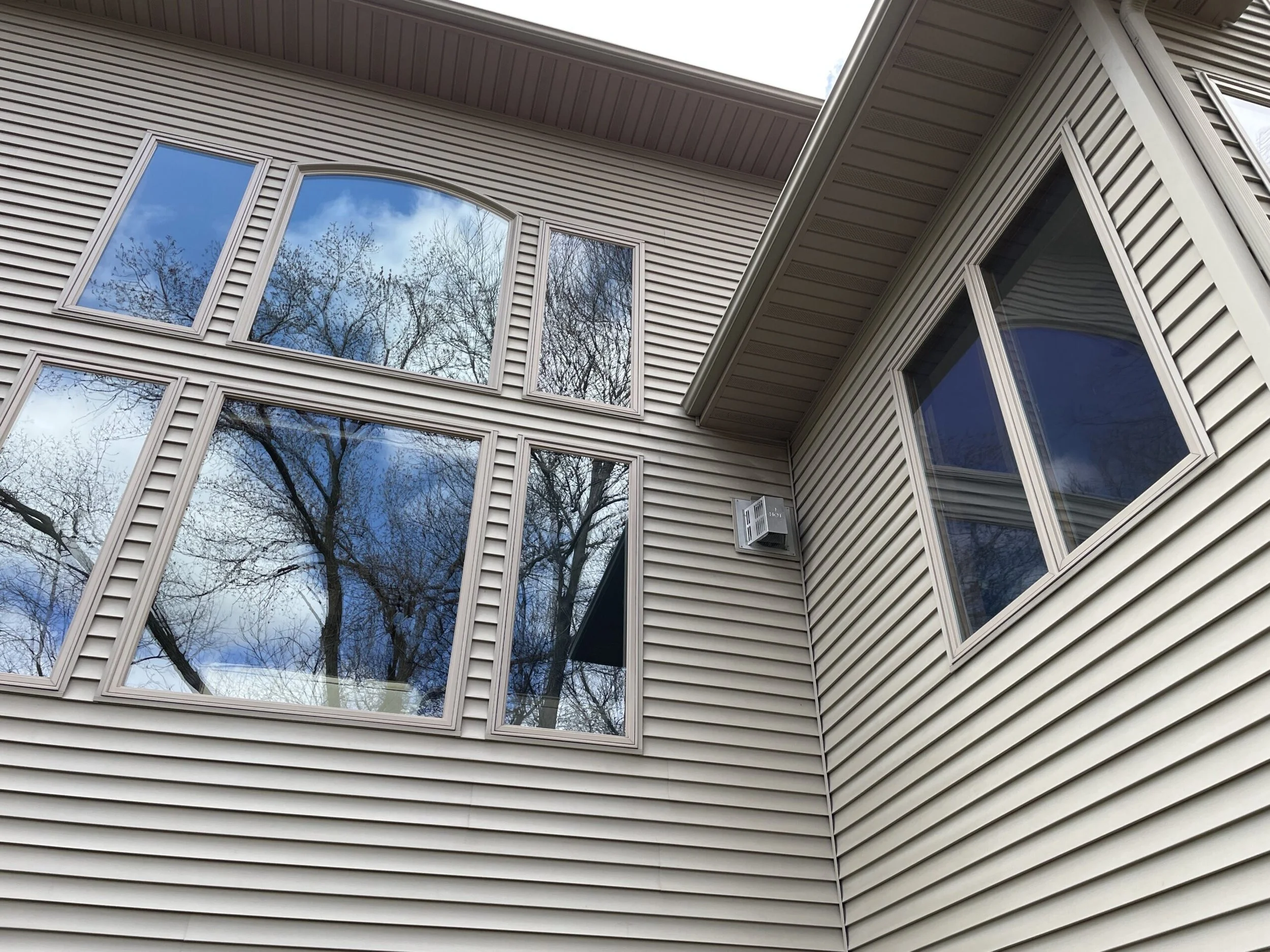 A close-up view of a beige residential house's exterior wall with multiple large windows that have been recently cleaned reflecting trees and sky, with beige siding and corner trim.