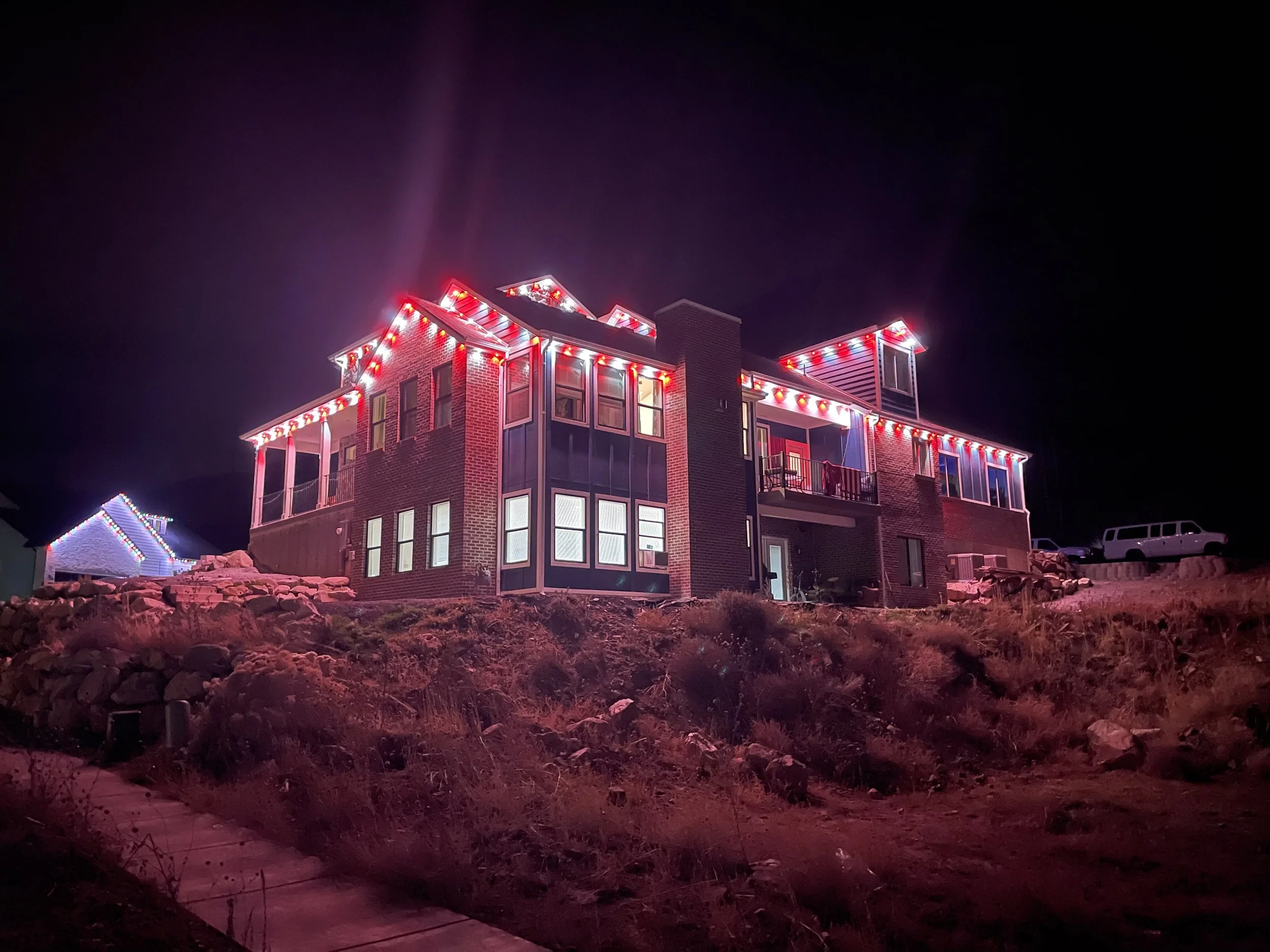 A multi-story brick house decorated with multicolored Christmas lights at night, with a rocky front yard and a sidewalk leading up to it.