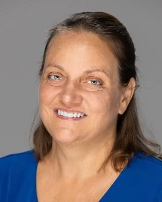 A woman with light skin, blue eyes, and brown hair smiling at the camera, wearing a blue top against a gray background.