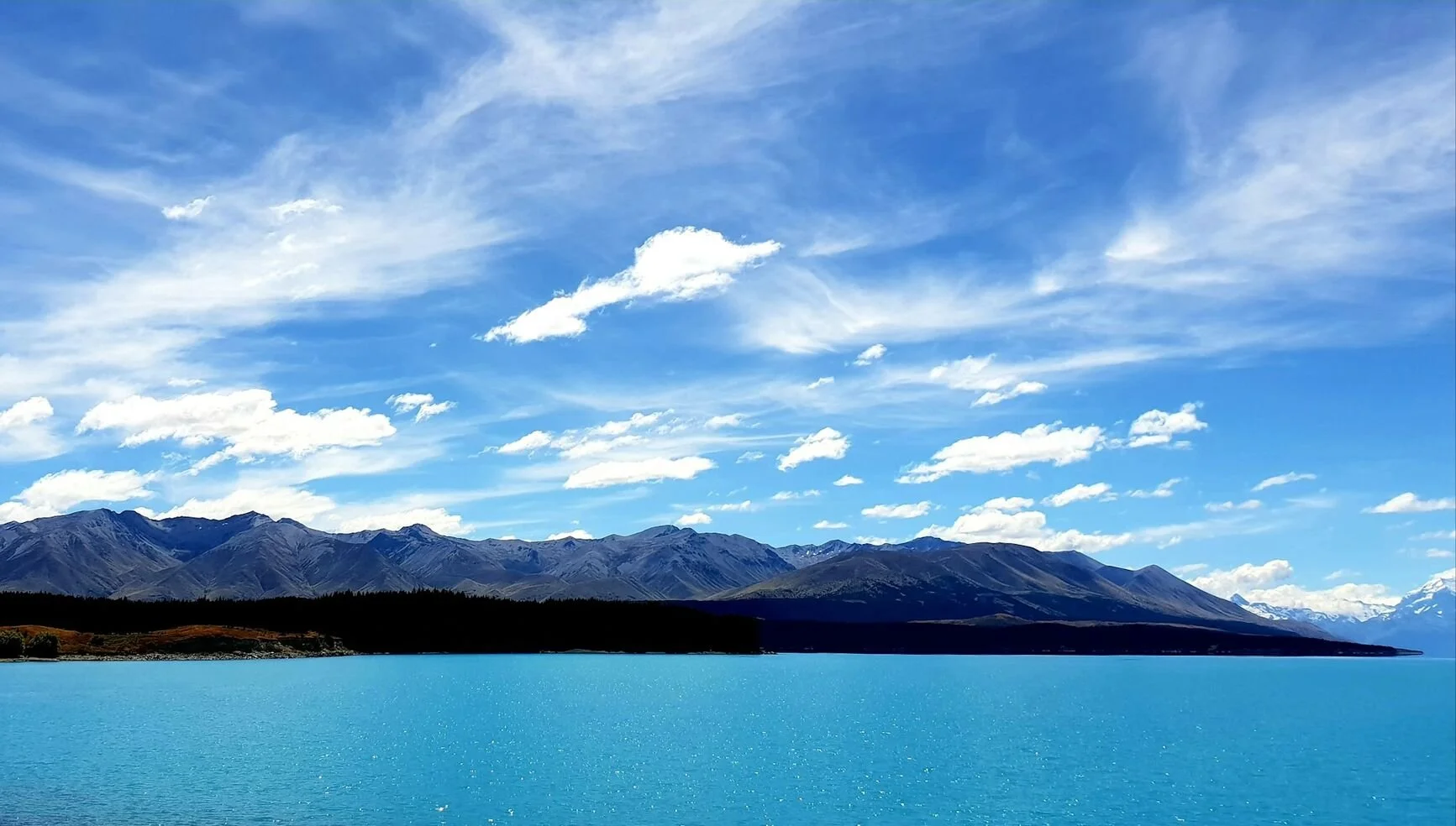 A landscape scene featuring a large blue lake, distant mountains, and a cloudy sky with wispy clouds.