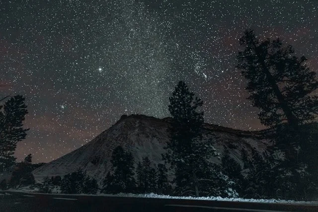 Night sky filled with stars above a mountain and trees, with a faint glow at the base of the mountain.