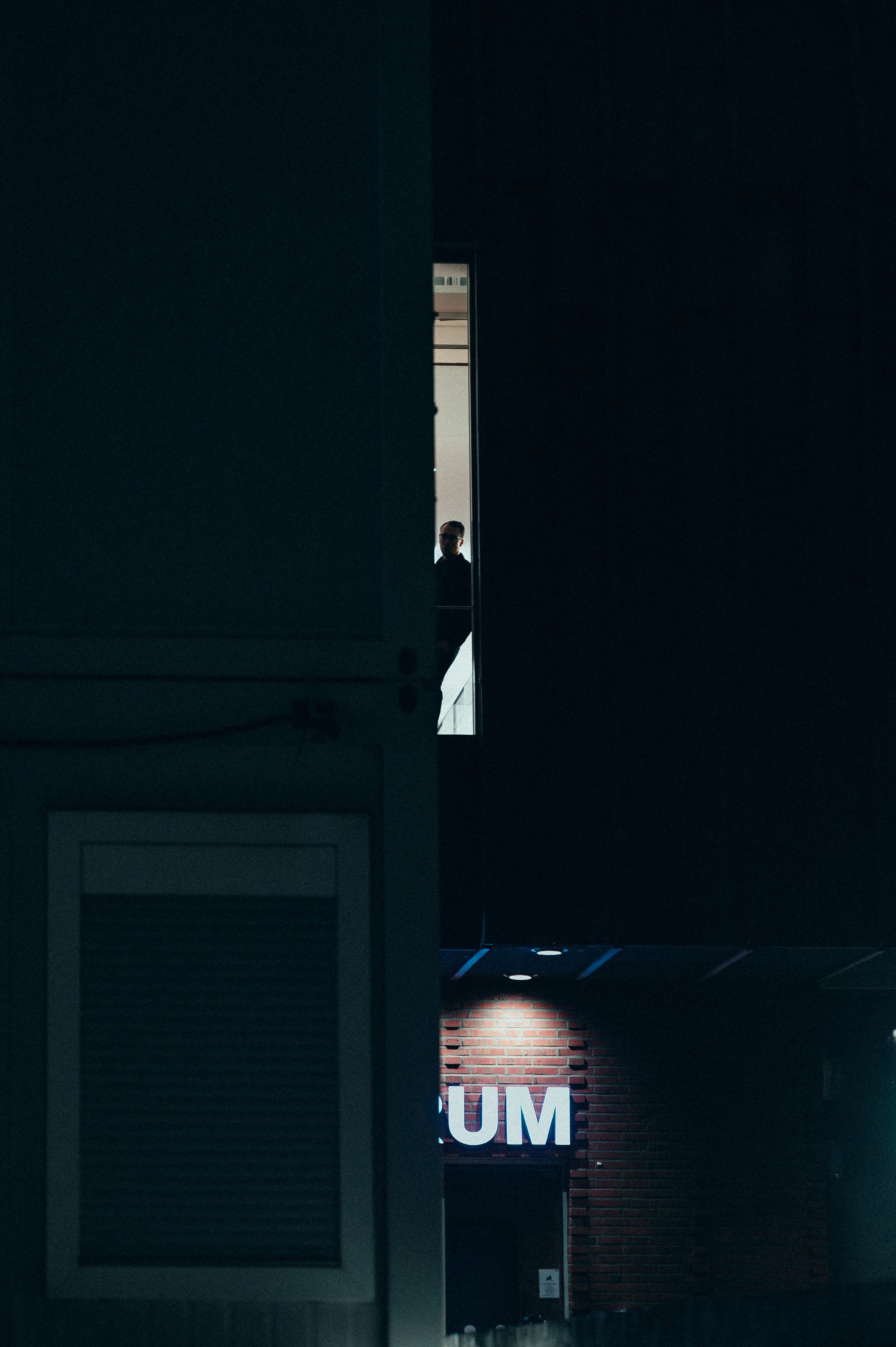 A dark, dimly lit urban scene at night showing a person standing on a balcony or ledge, looking out from behind a narrow opening in a building, with a brightly lit sign reading 'UM' on a brick wall below.