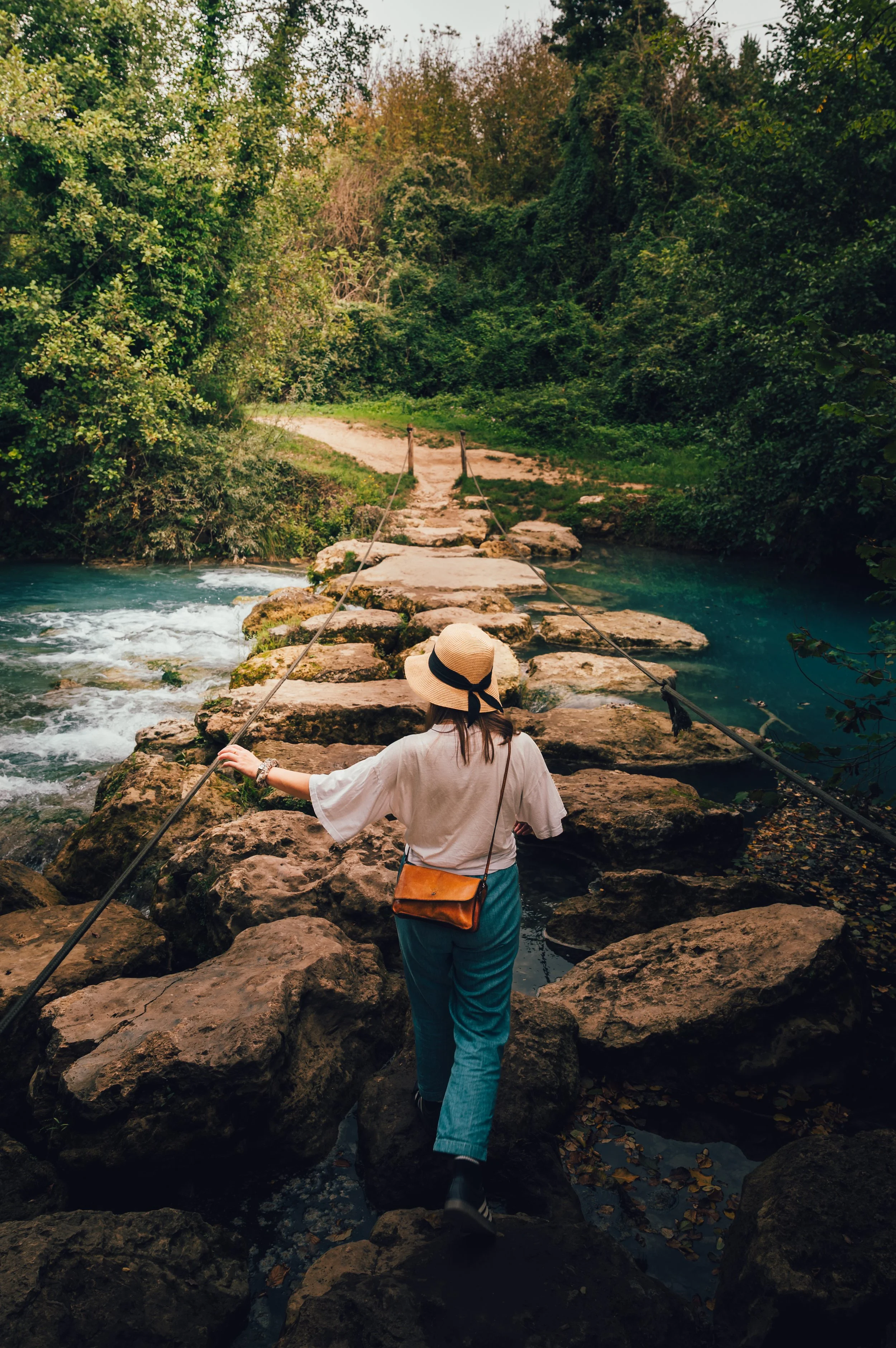 A woman crossing a rocky footbridge over a river in a lush, wooded area during daytime.