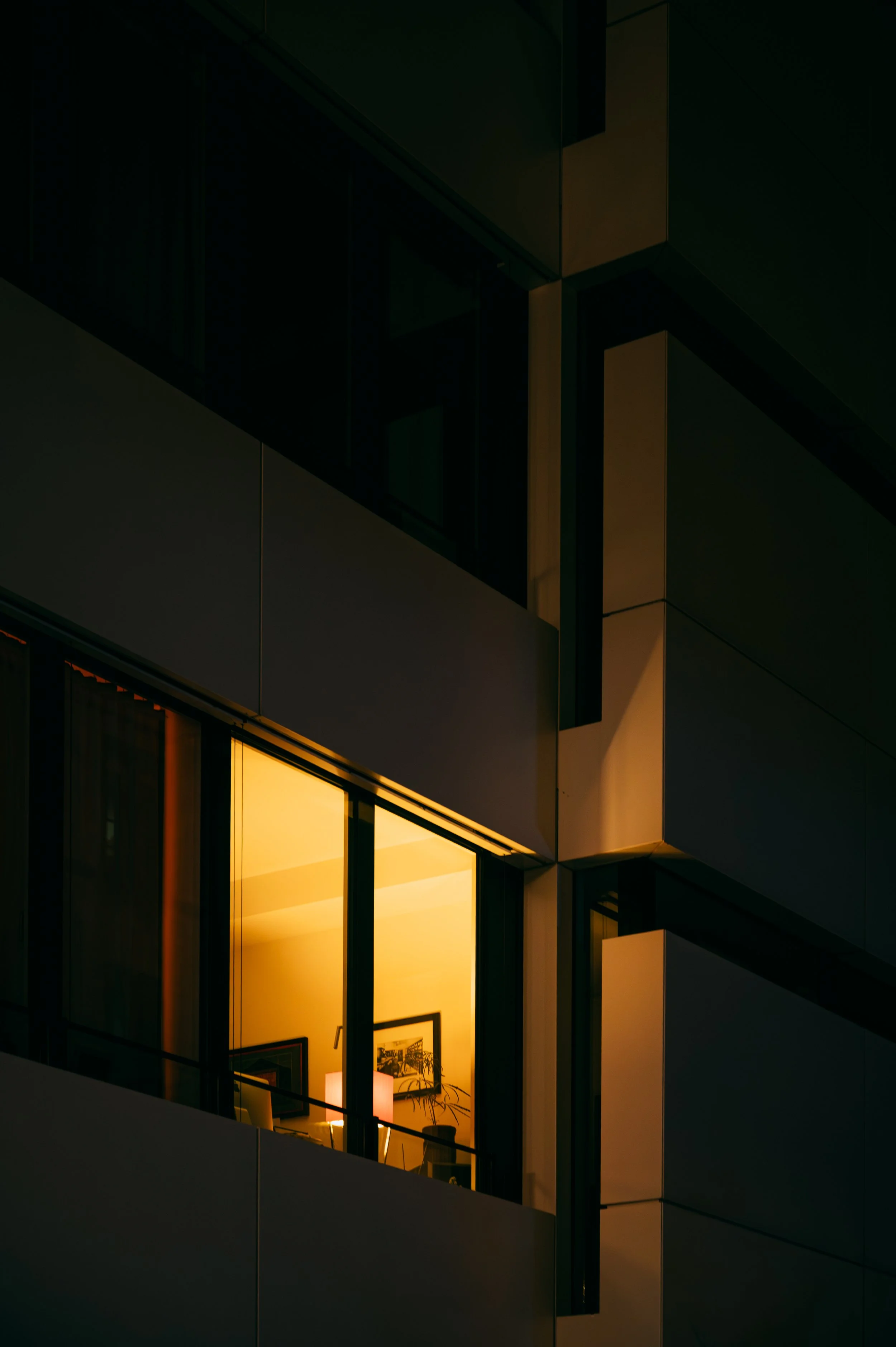 Nighttime view of modern apartment building with lit interior seen through large windows, showing framed pictures, houseplants, and a table lamp.