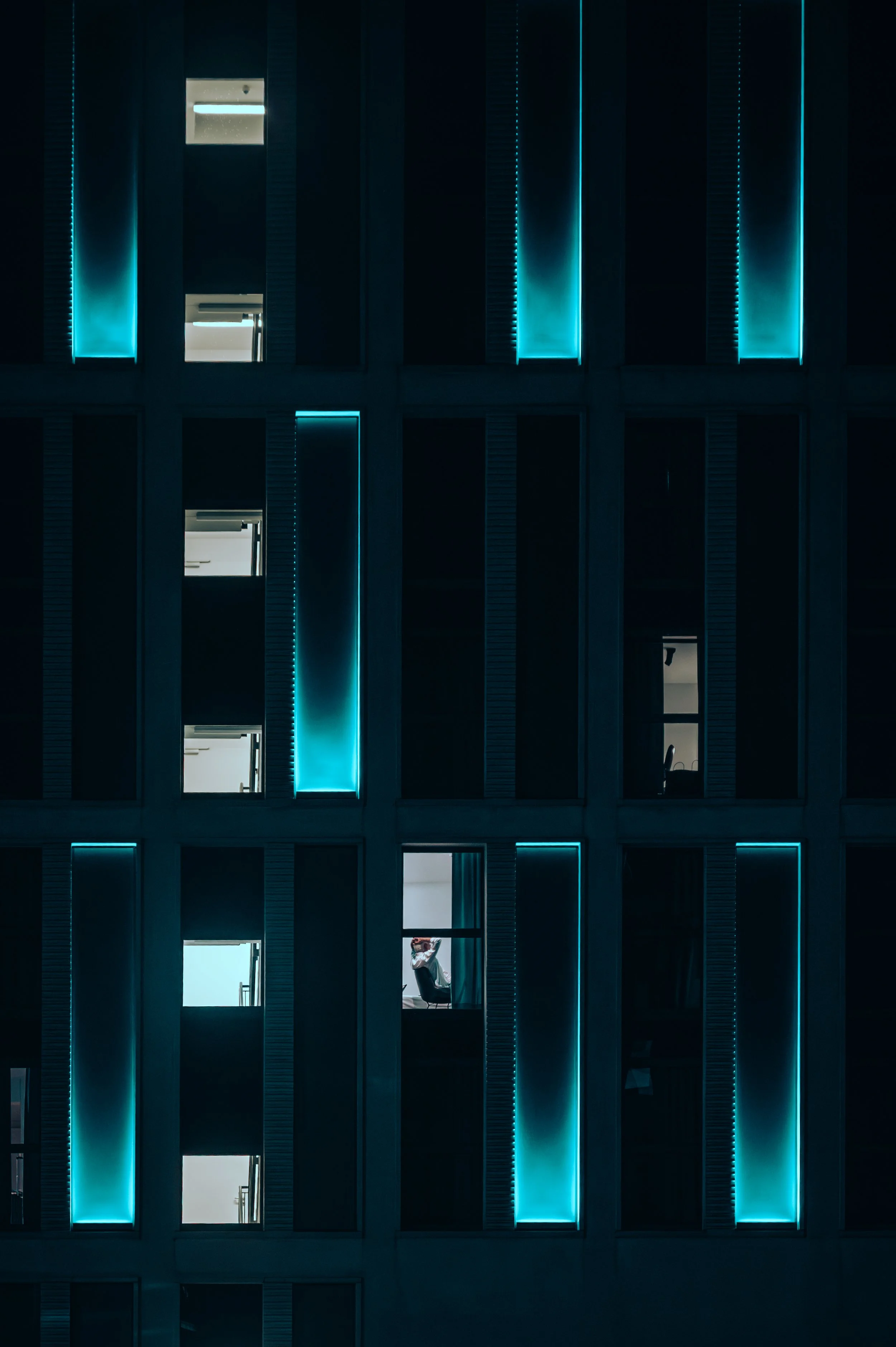Night view of a modern building with illuminated vertical blue neon lights in the windows and a person visible through one window working at a desk.