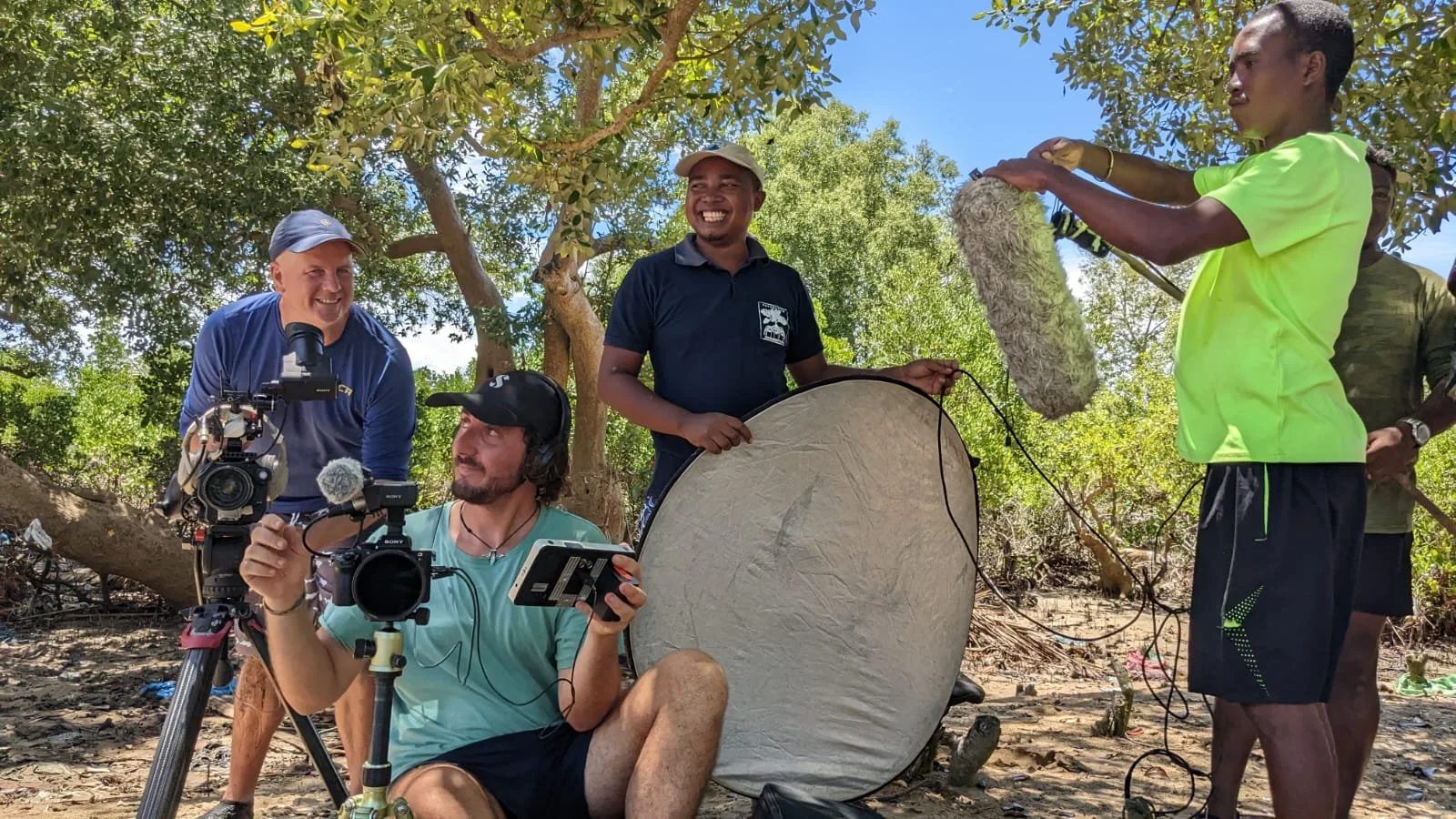 Filmmakers and crew filming outdoors in a forest, with bright sunlight, trees, and equipment such as cameras, microphones, and reflector, capturing a scene involving local people.