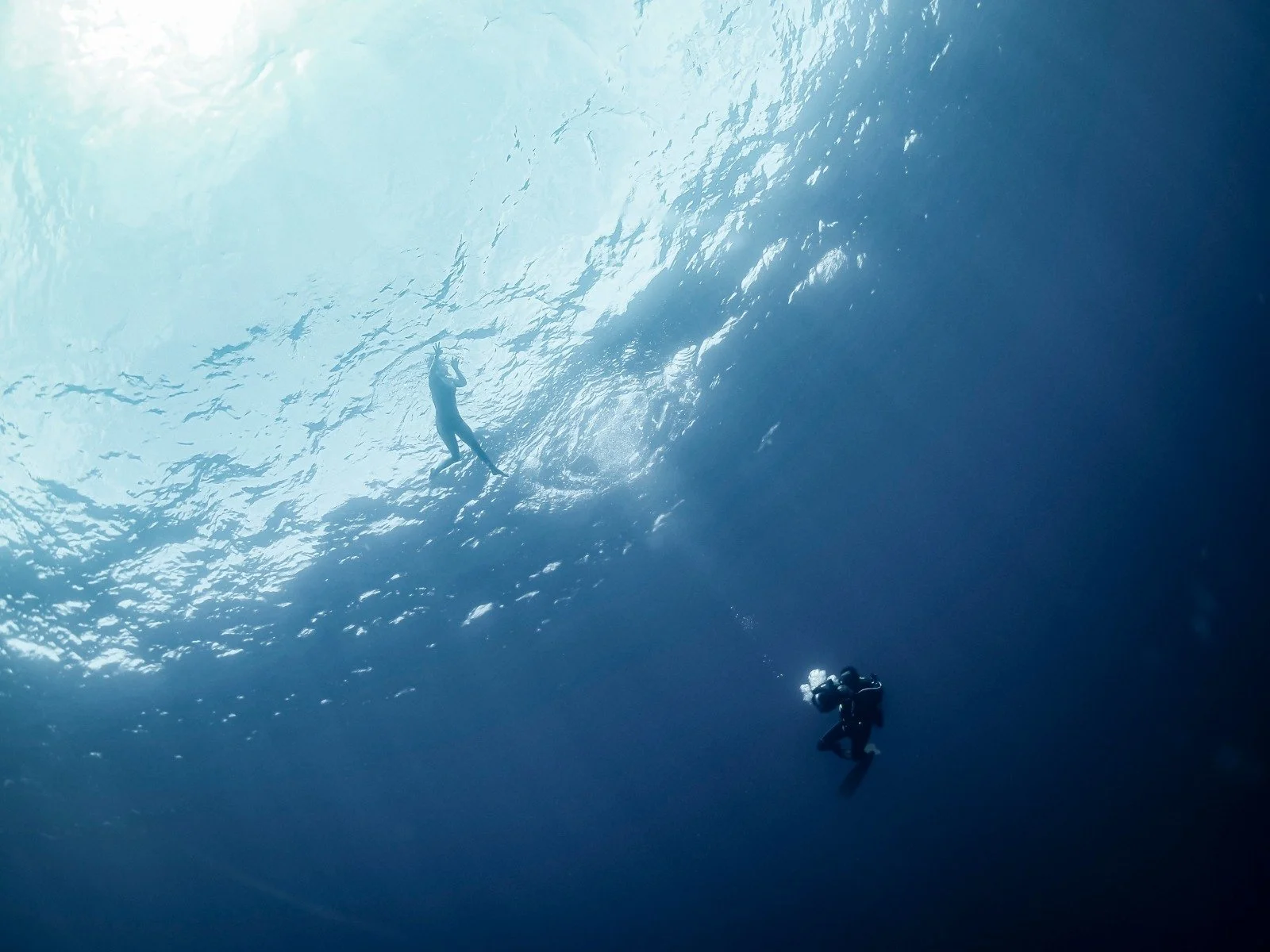 Two scuba divers underwater, one near the surface and the other deeper in the ocean, with sunlight filtering through the water.