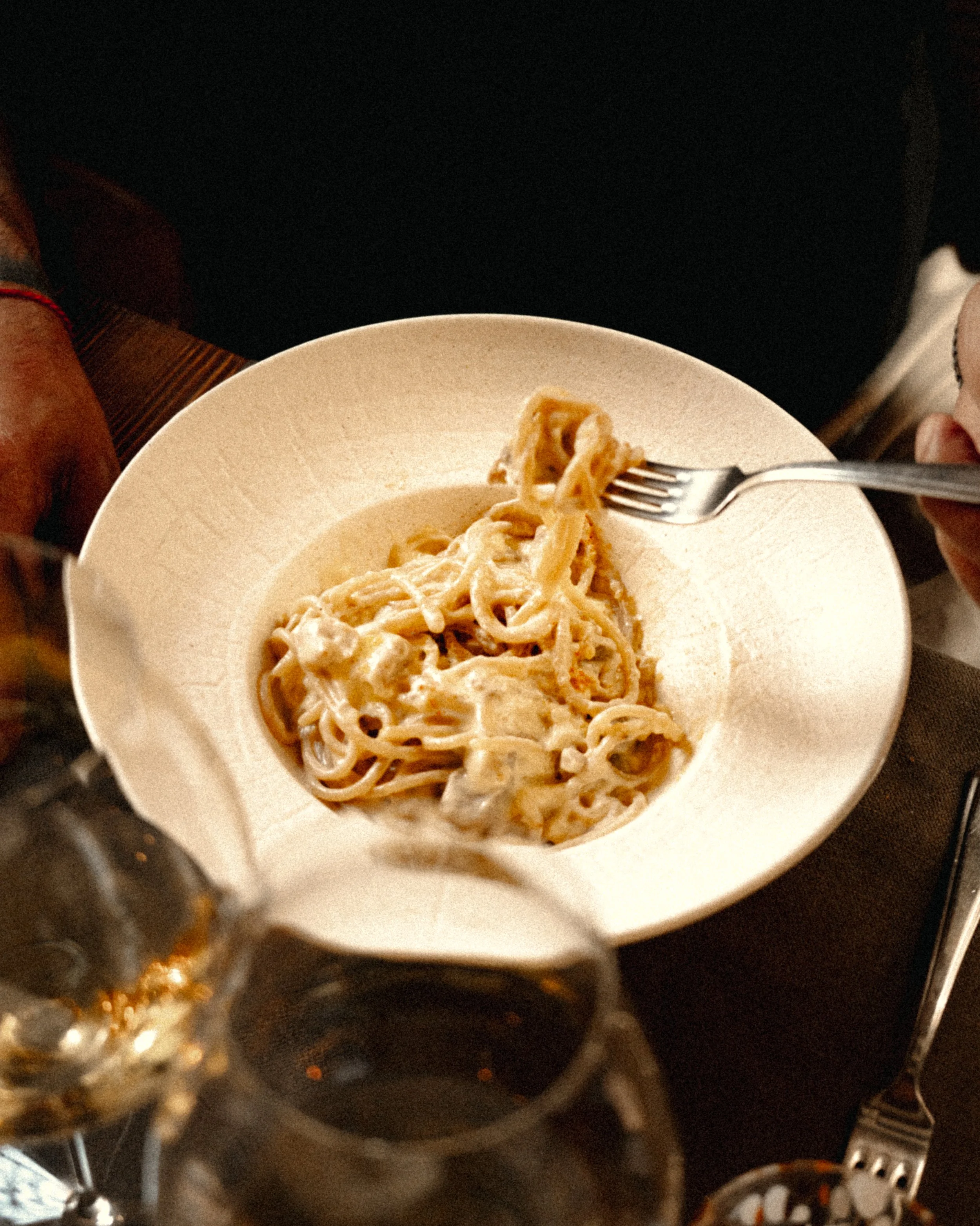 A bowl of creamy fettuccine pasta with pieces of chicken, served on a dark table with wine glasses in the foreground.