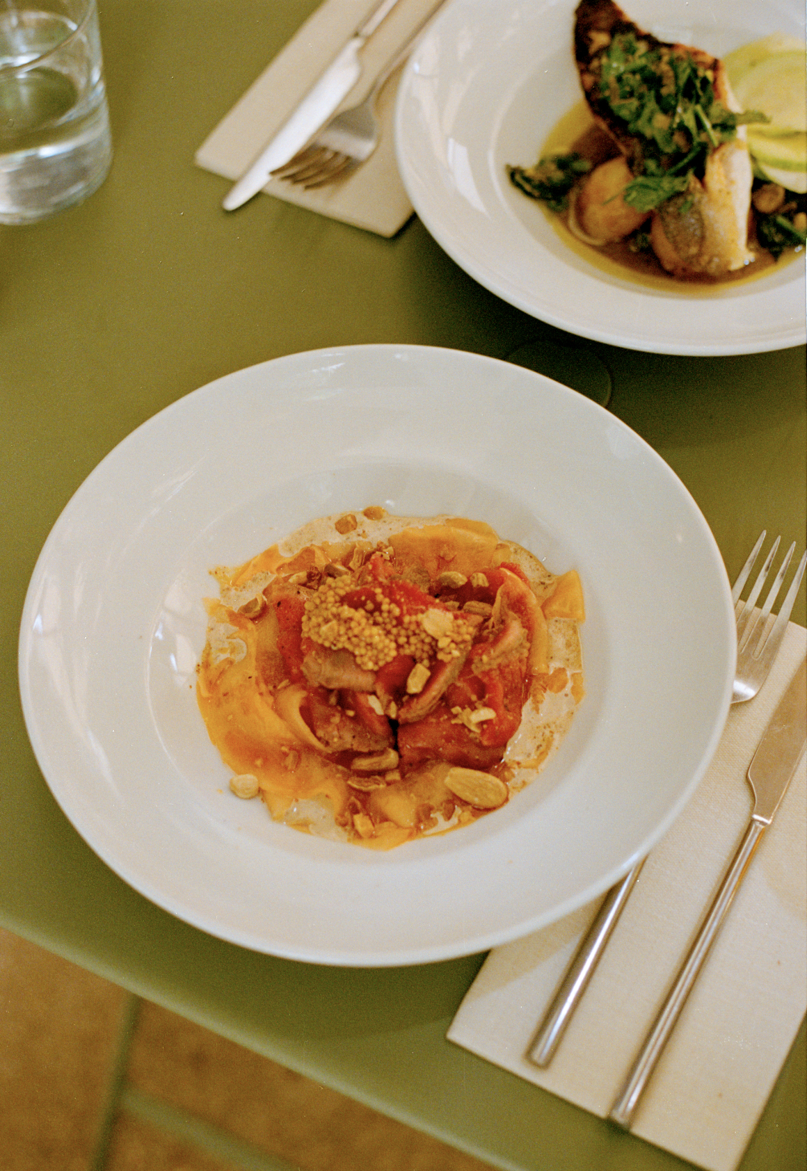 A plate of Italian pasta with tomato sauce, topped with nuts, on a green table, with a glass of water and another dish in the background.