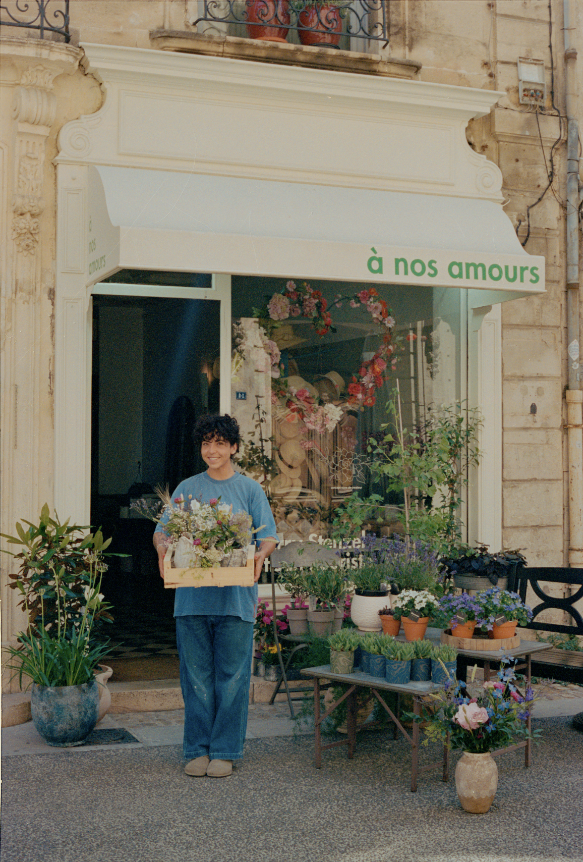 A woman standing outside a flower shop holding a wooden basket of flowers. The shop has a white awning with green text "à nos amours". There are potted plants and flowers displayed outside the shop, with some inside as well visible through the large window.