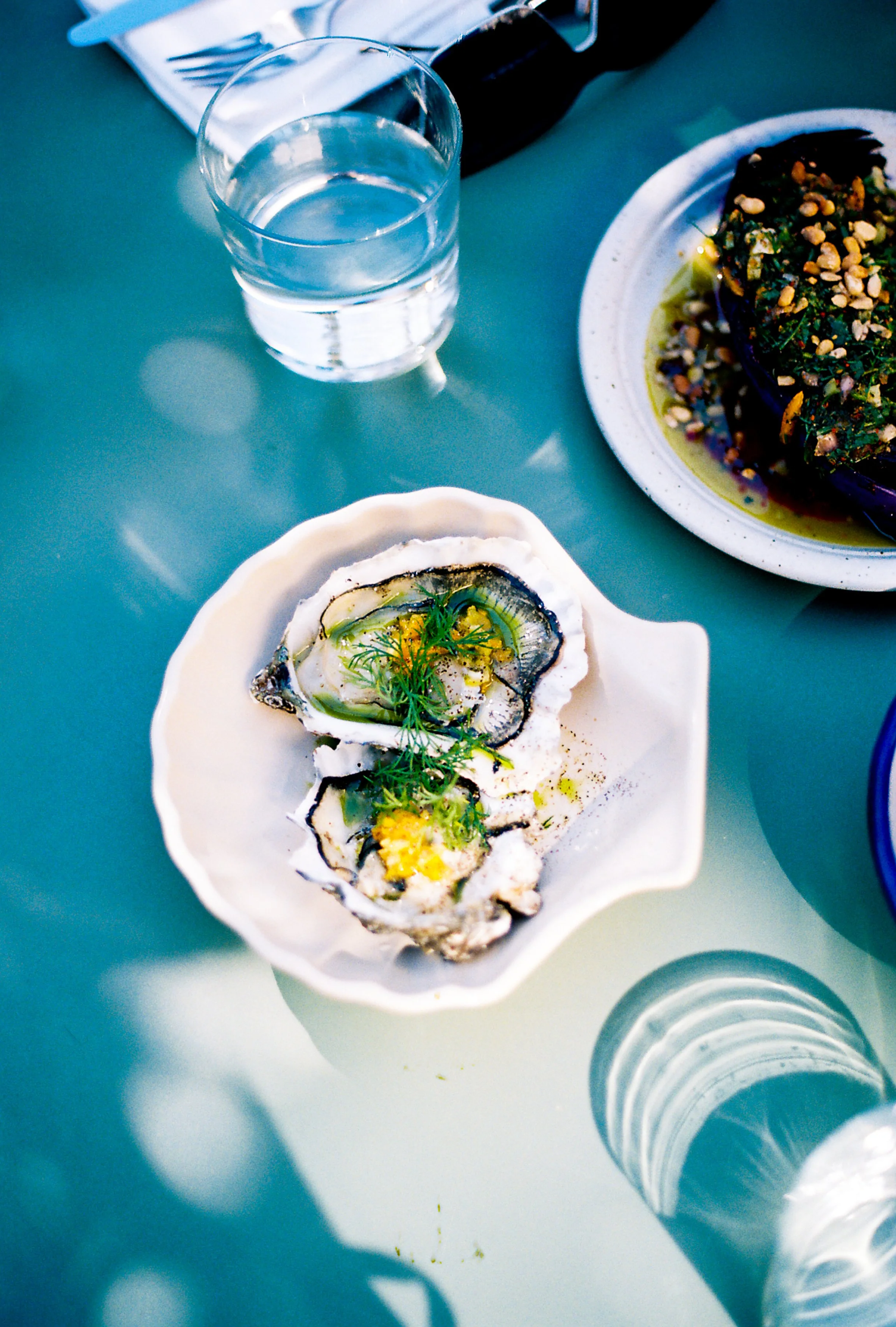 A white dish with two oysters garnished with herbs and lemon slices on a light green surface. Nearby are glasses of water and other plates of food.