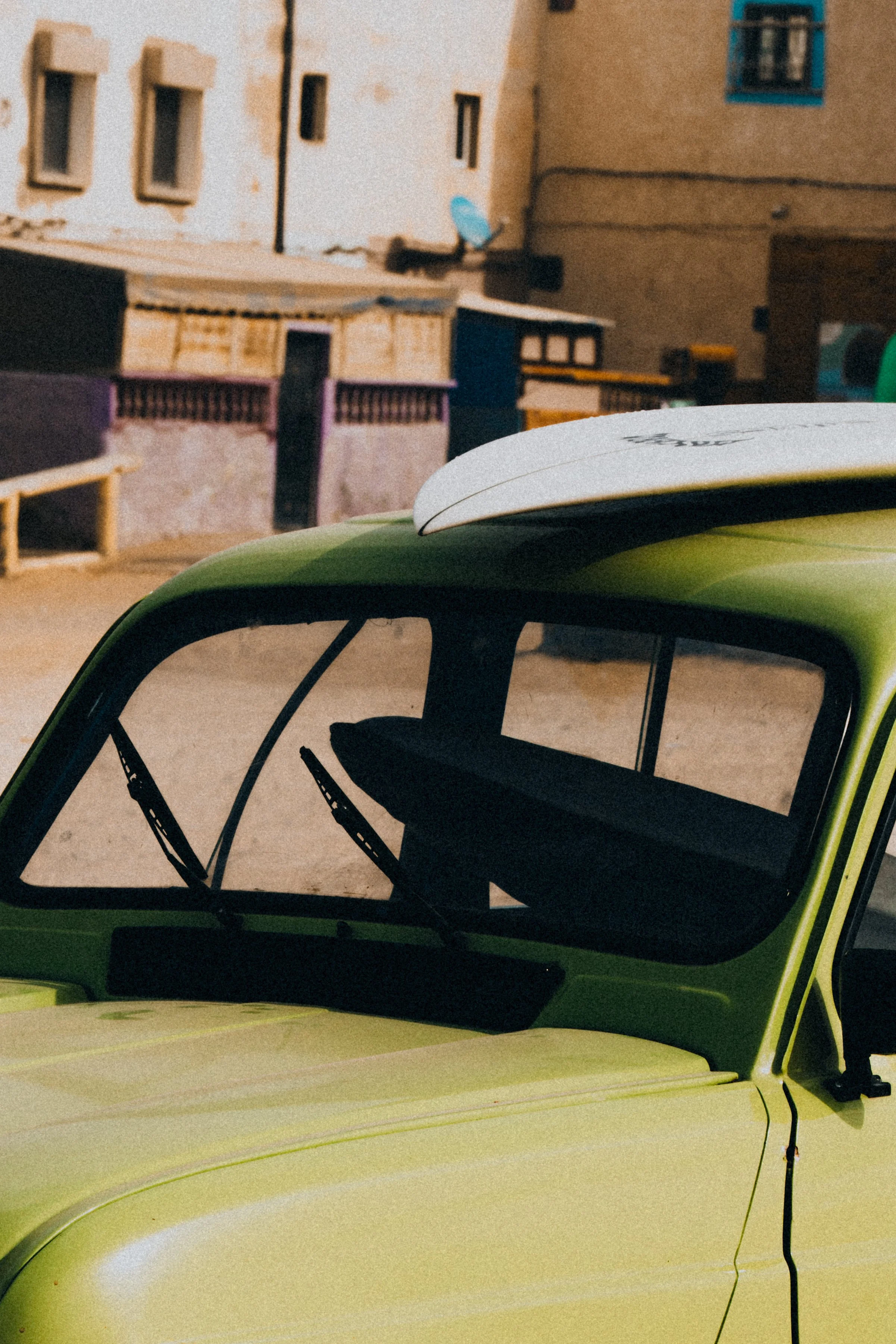 Close-up of a bright green vintage car with a white spoiler, parked outdoors near a rustic building with small windows and a satellite dish.