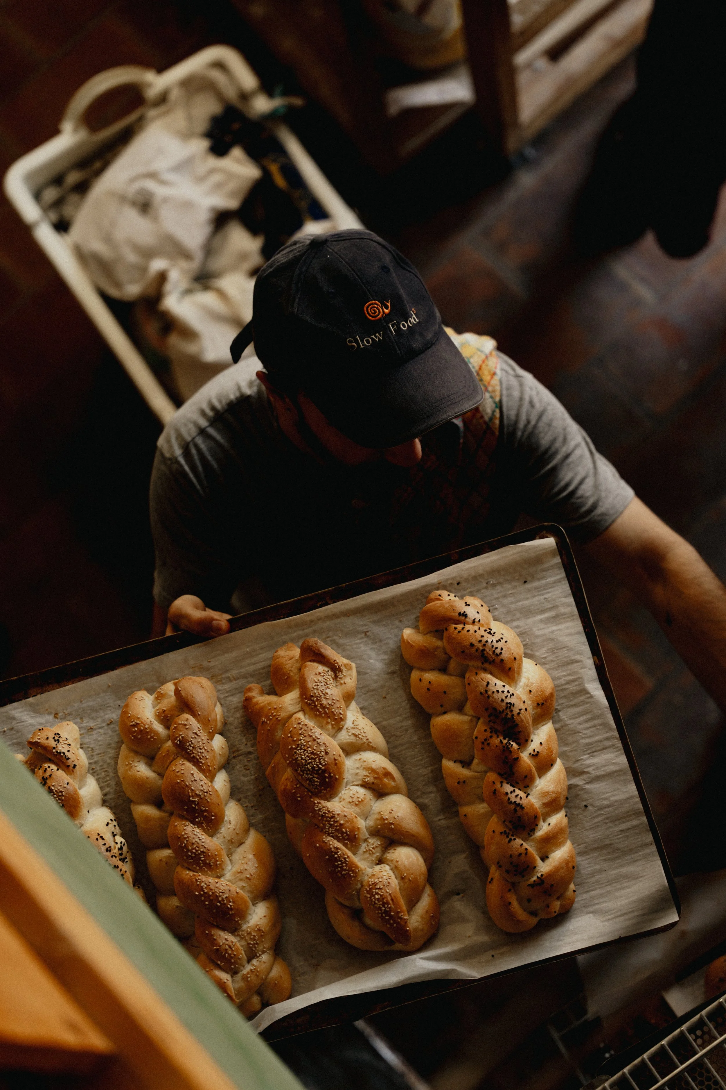 A person wearing a black cap with on it the words 'Slow Food' and a curled shell logo, stands above a tray of freshly baked twisted bread loaves topped with sesame and black seeds, seen from above.