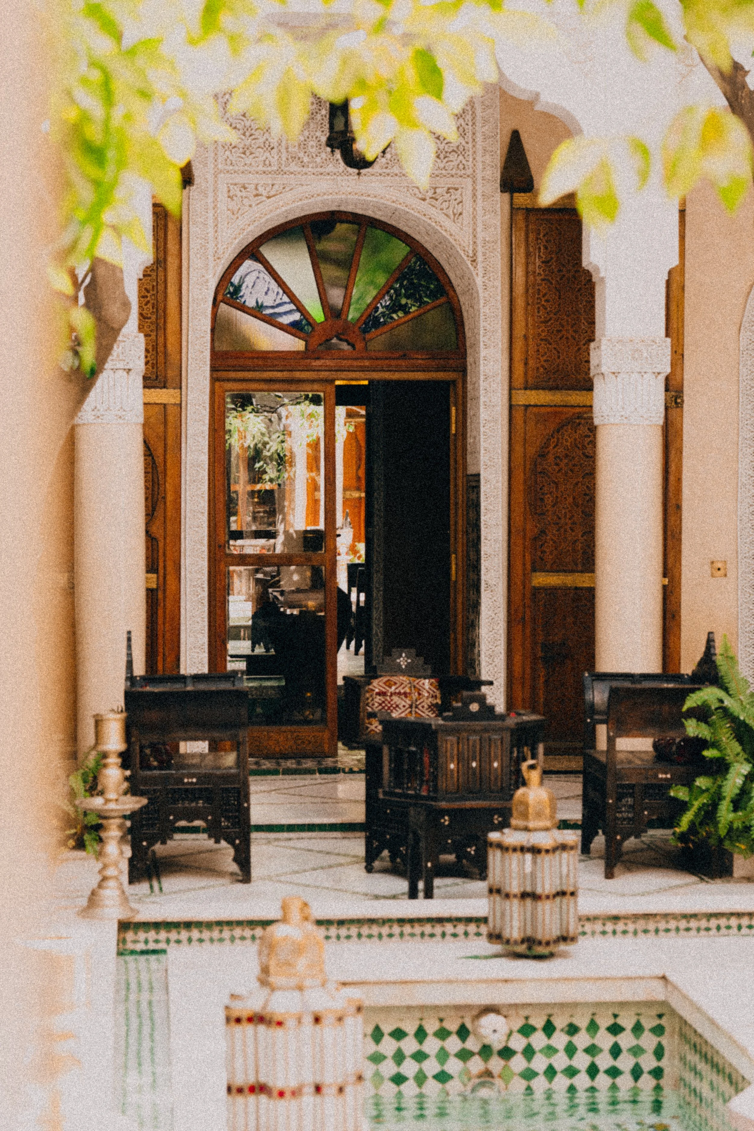 An ornate entrance to a building with a wooden door and arched window, flanked by columns, and decorated with intricate carvings, inside a courtyard with chairs, plants, and a small fountain.