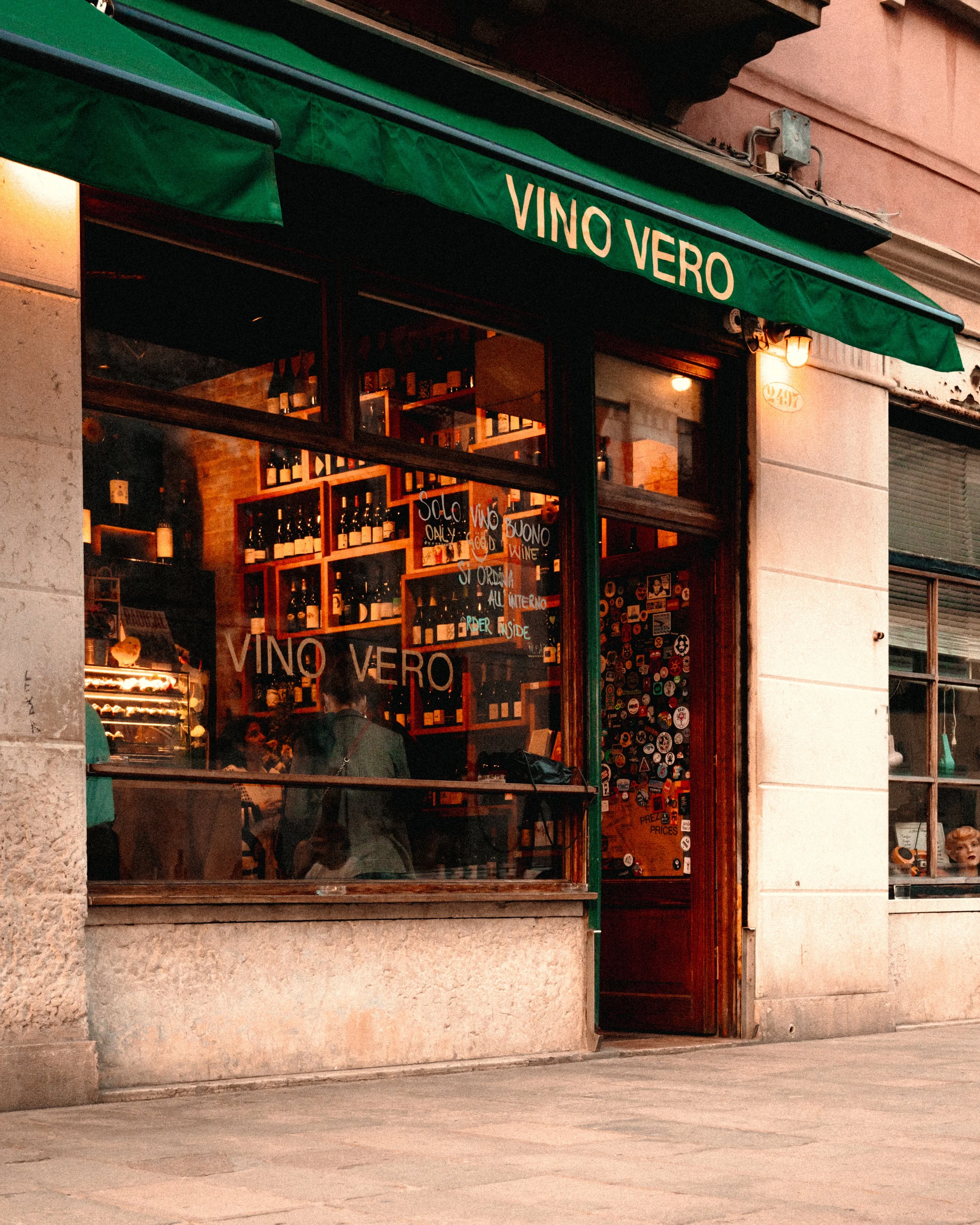 Exterior of a wine shop with a green awning labeled 'Vino Vero', showing a window display inside with wine bottles and decorative stickers on the door.