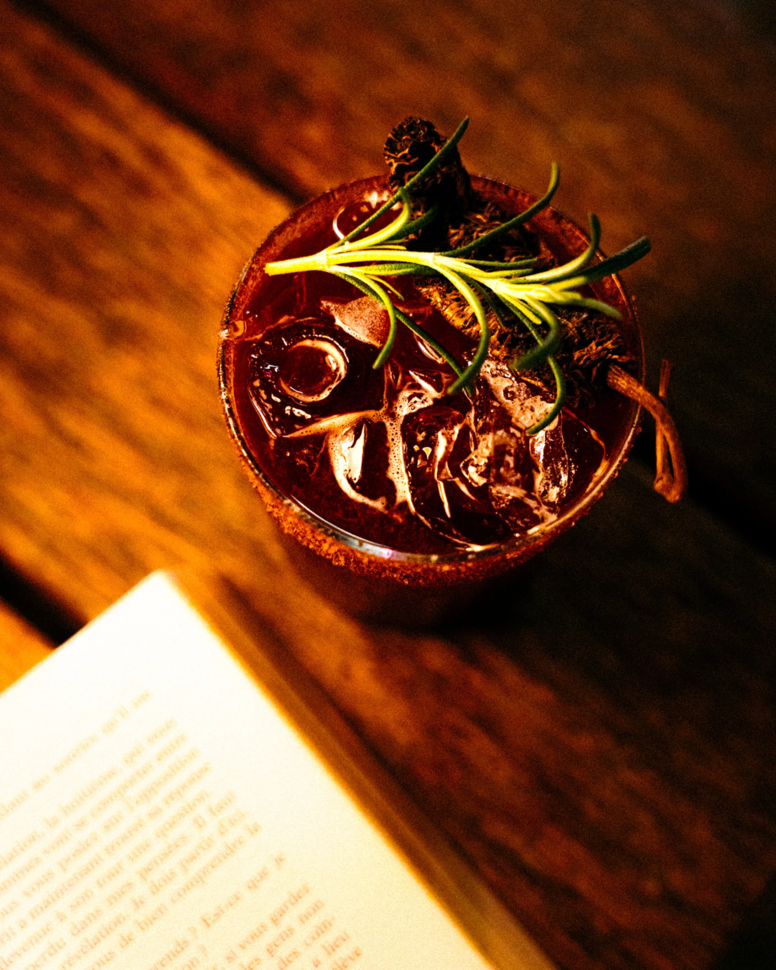 A glass of dark-colored cocktail garnished with a sprig of rosemary and a cinnamon stick, placed on a wooden surface near a book.