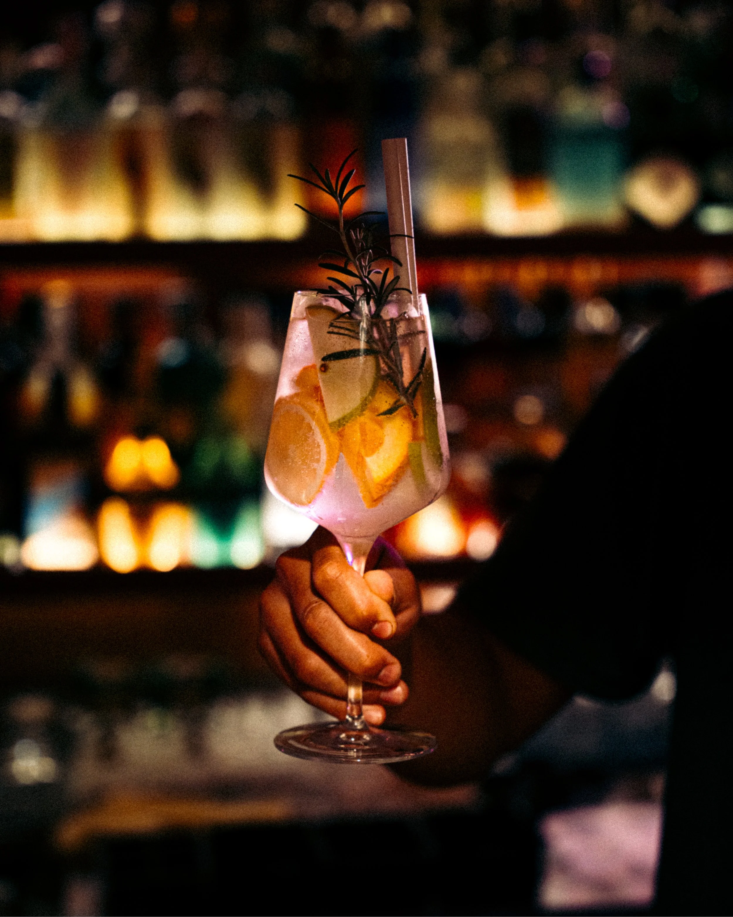 Hand holding a tall glass of cocktail with lemon and orange slices and a sprig of rosemary, in a dimly lit bar.