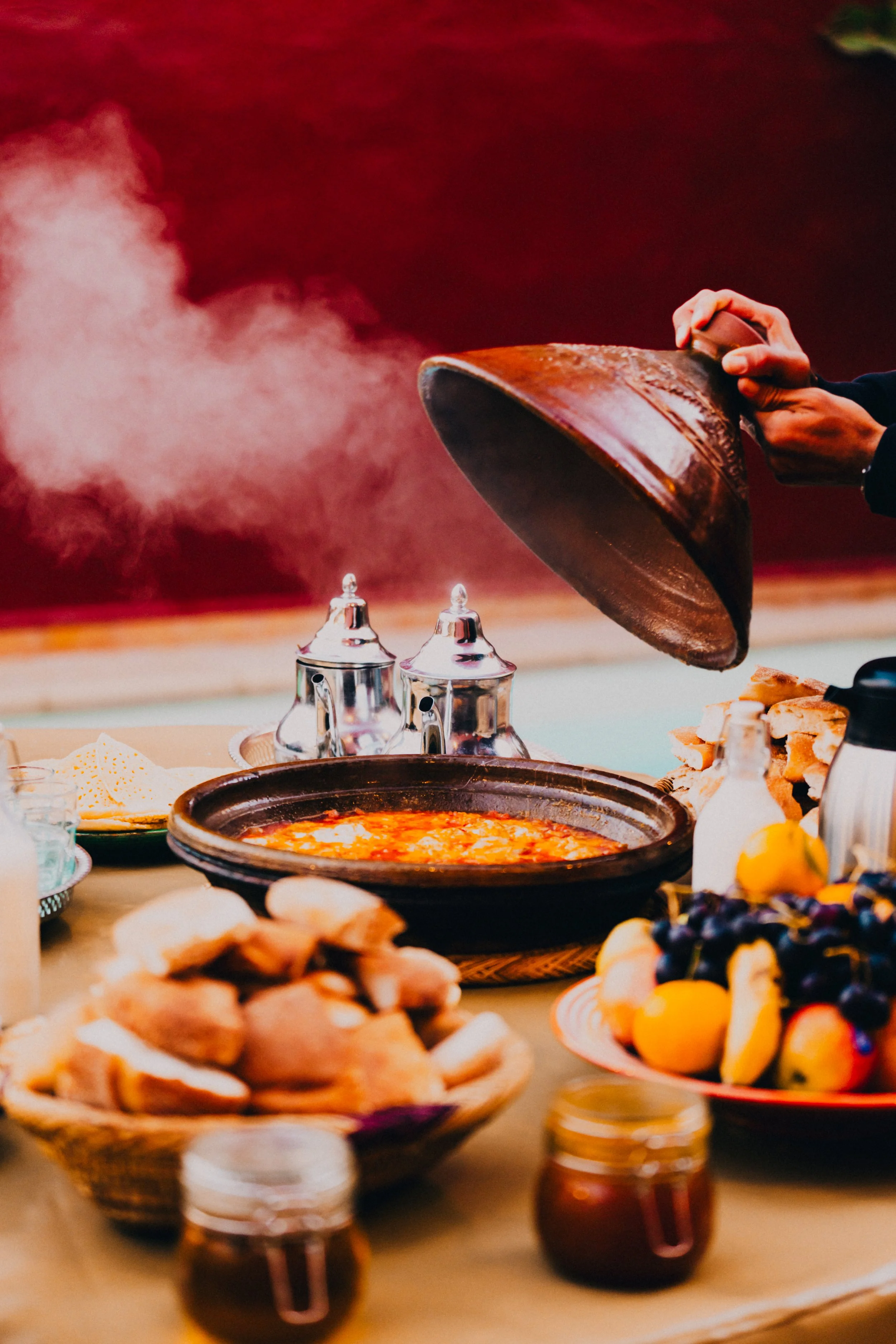 Person pouring soup into a bowl at a table with bread, fruit, and condiments, on a red background.