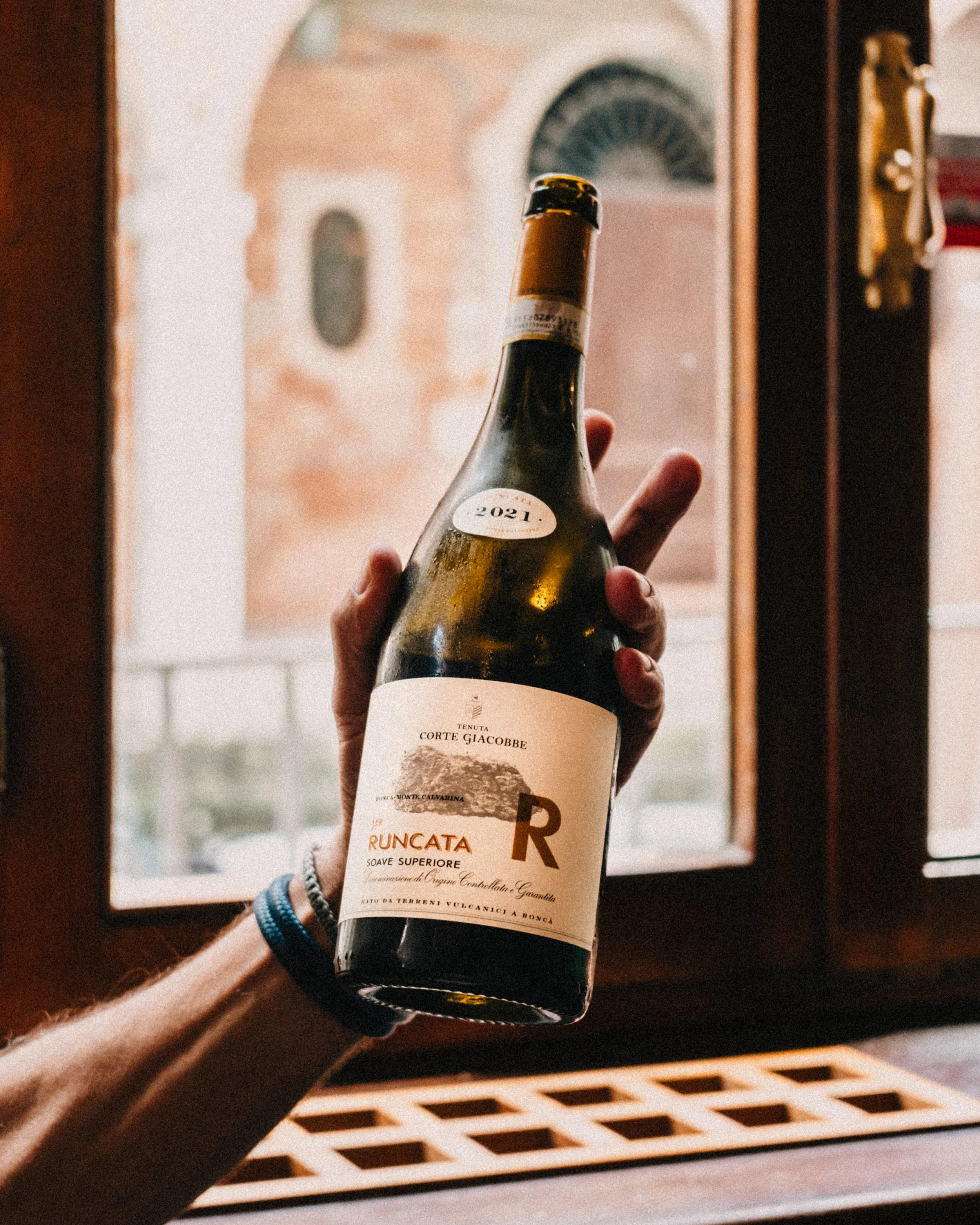 A person holding a bottle of Runcata wine inside a room with a window showing an outdoor brick building.