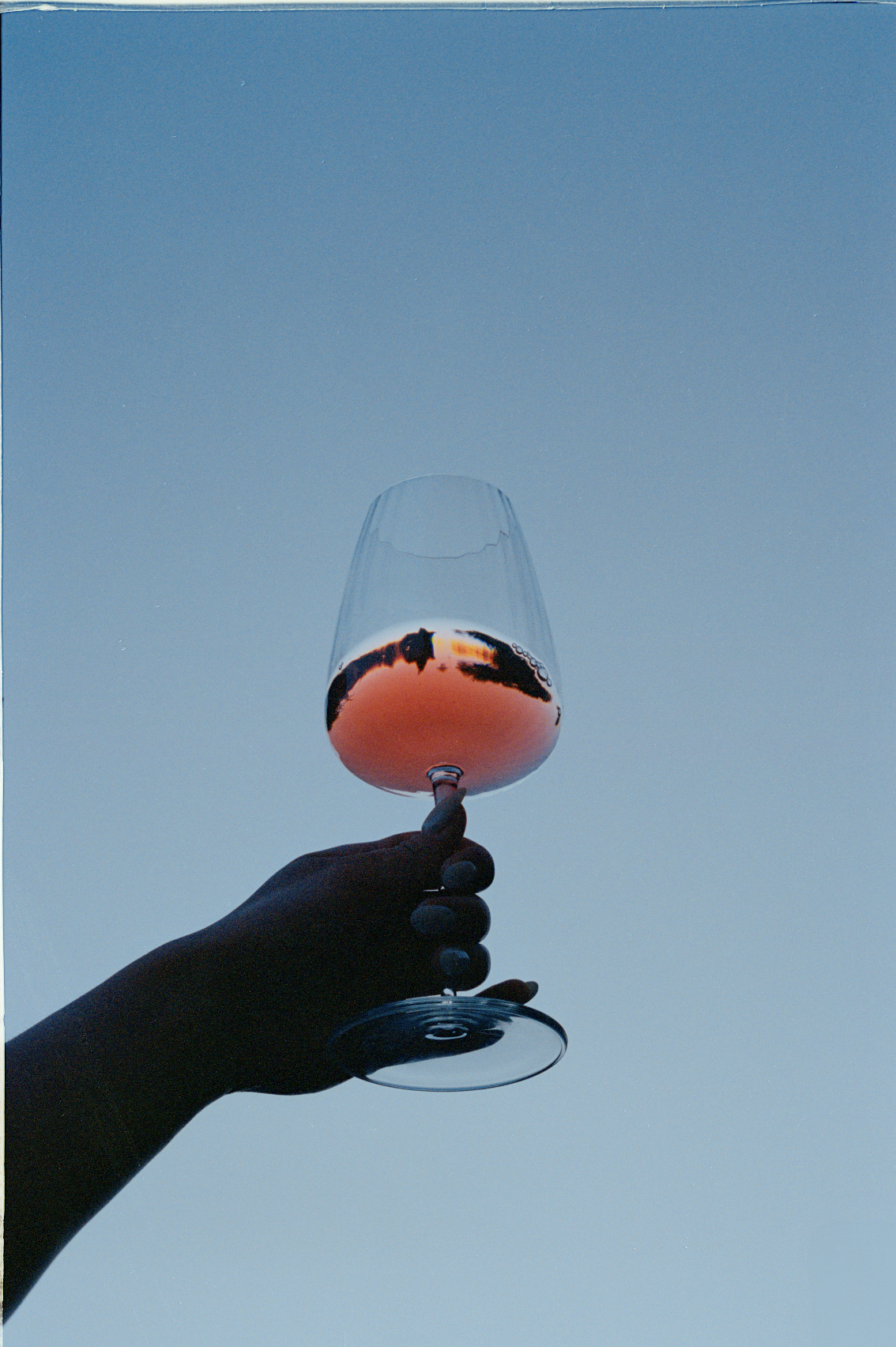 A person with black painted nails holding a glass of rosé wine against a clear blue sky