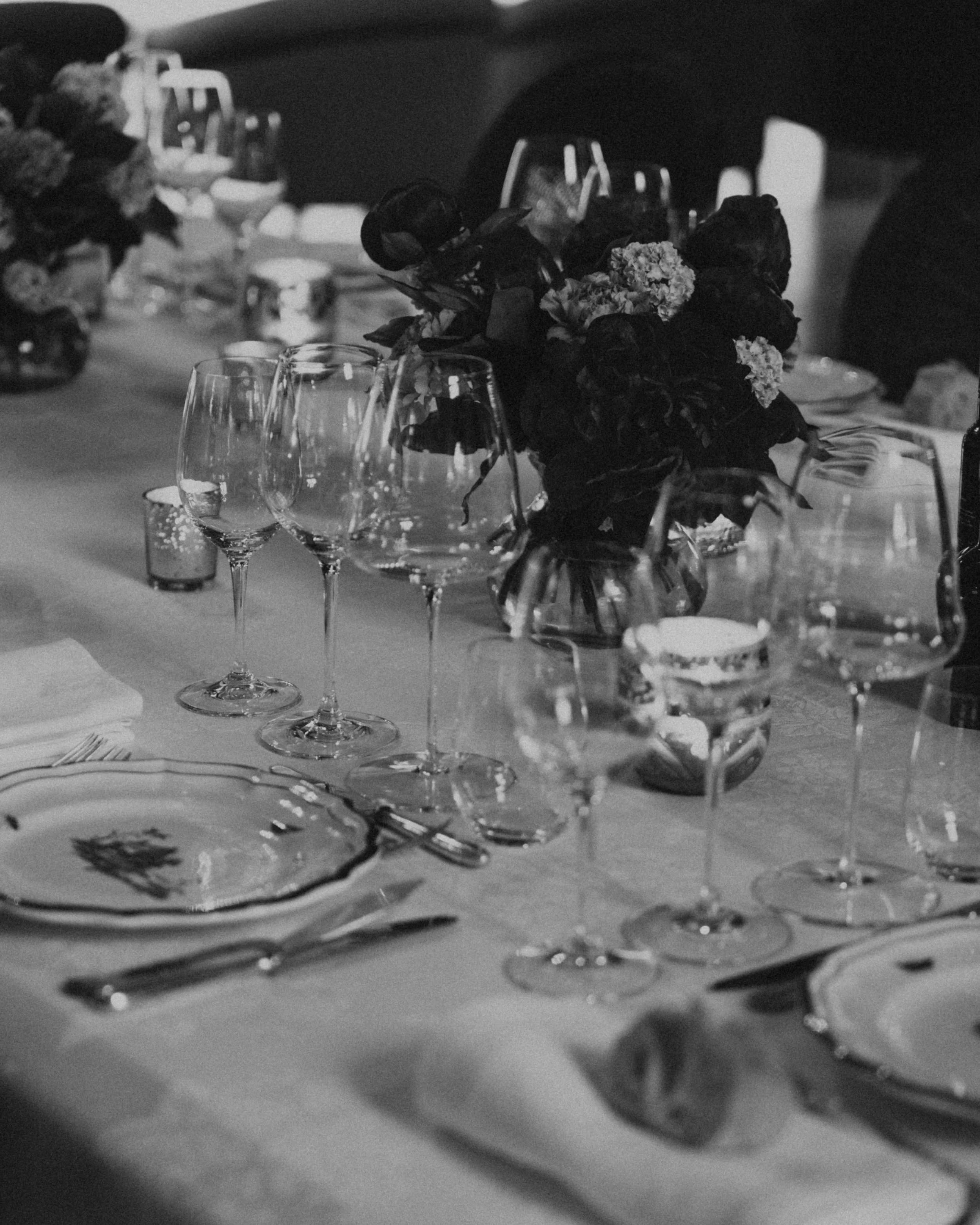 A formal dining table set with multiple wine glasses, plates, silverware, and floral centerpieces in a dimly lit setting.