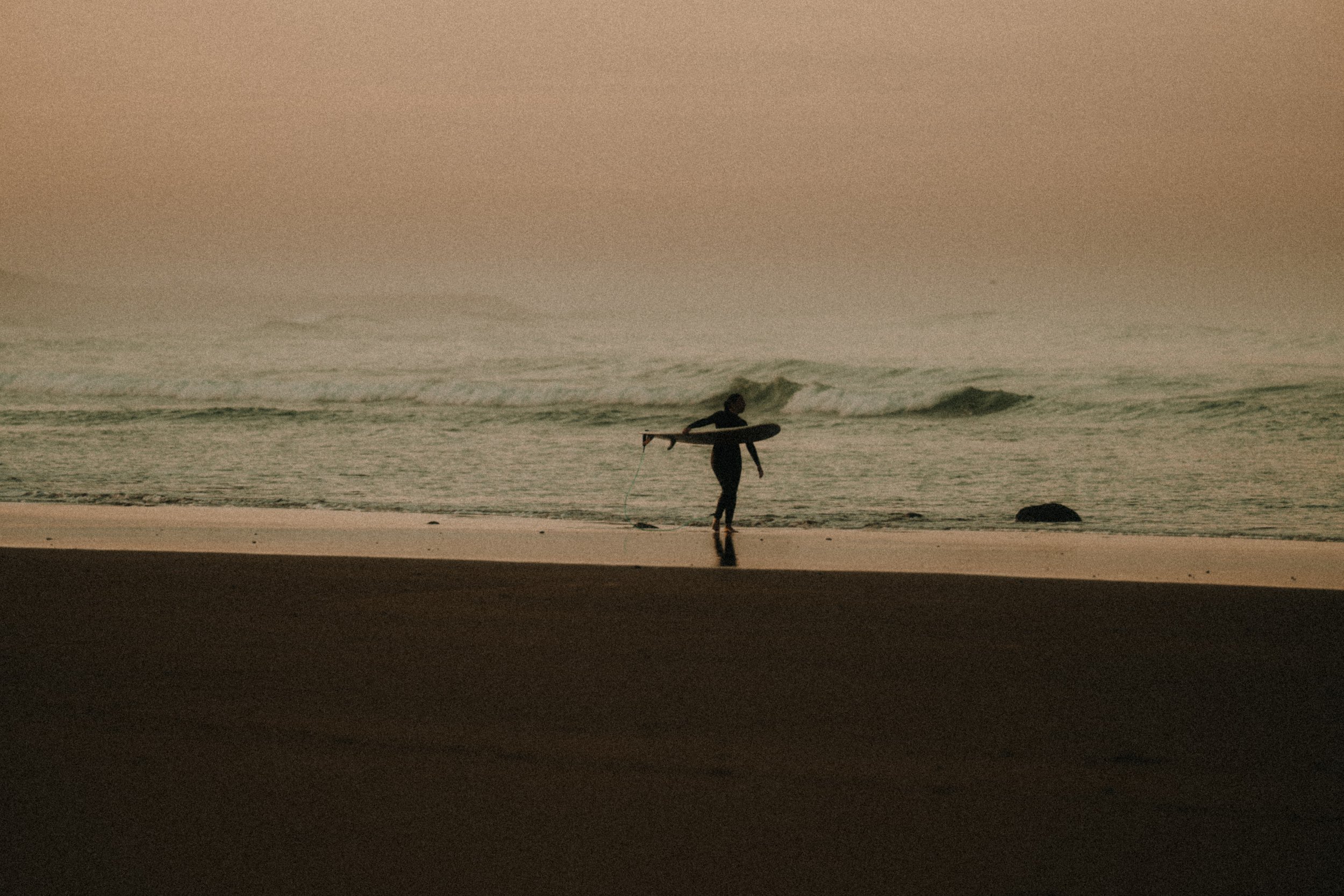 A person walking on the beach with a surfboard at sunset or sunrise, with waves in the background.