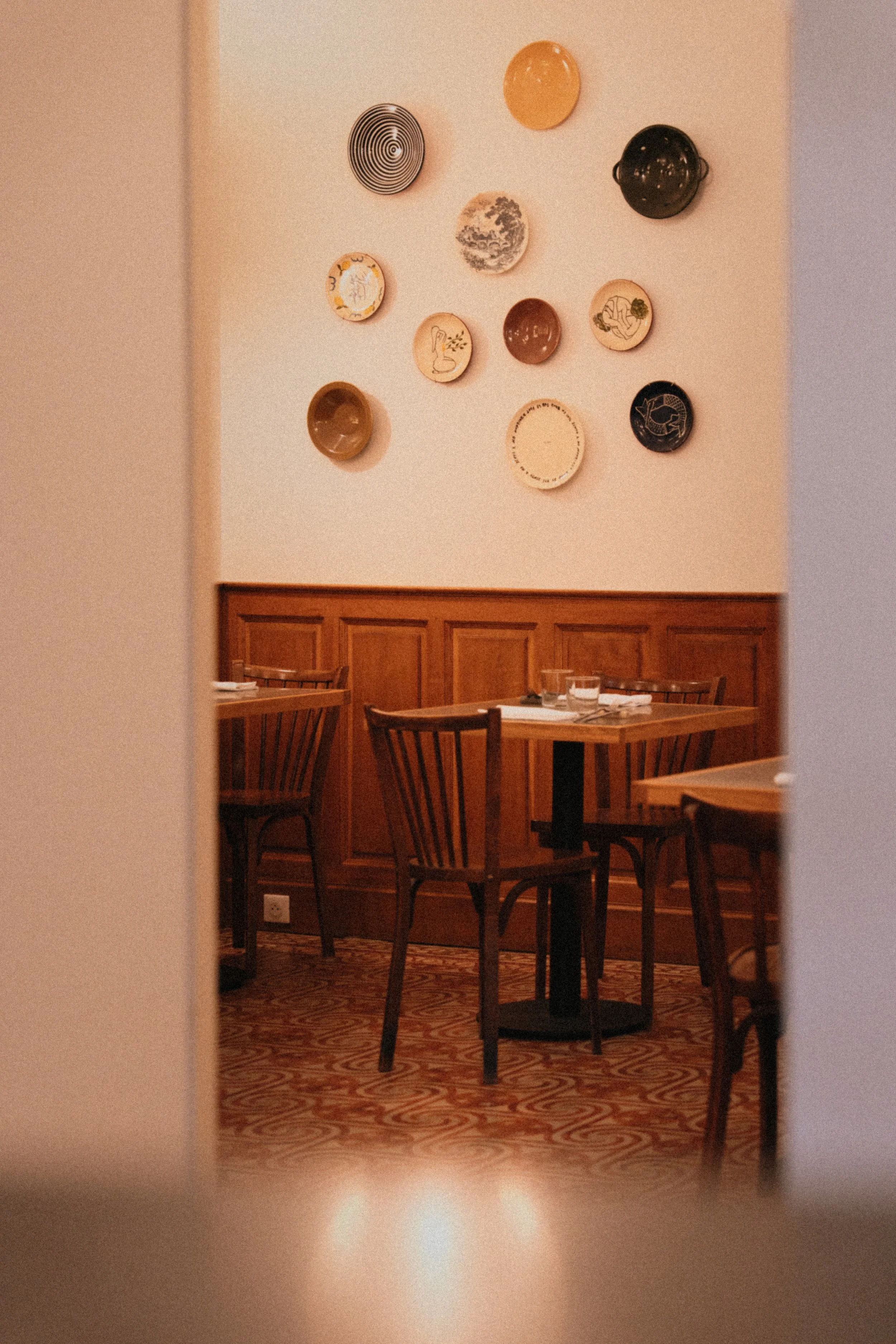 A cozy restaurant with wooden tables and chairs, decor featuring decorative plates and artwork on the wall, and a patterned carpet.