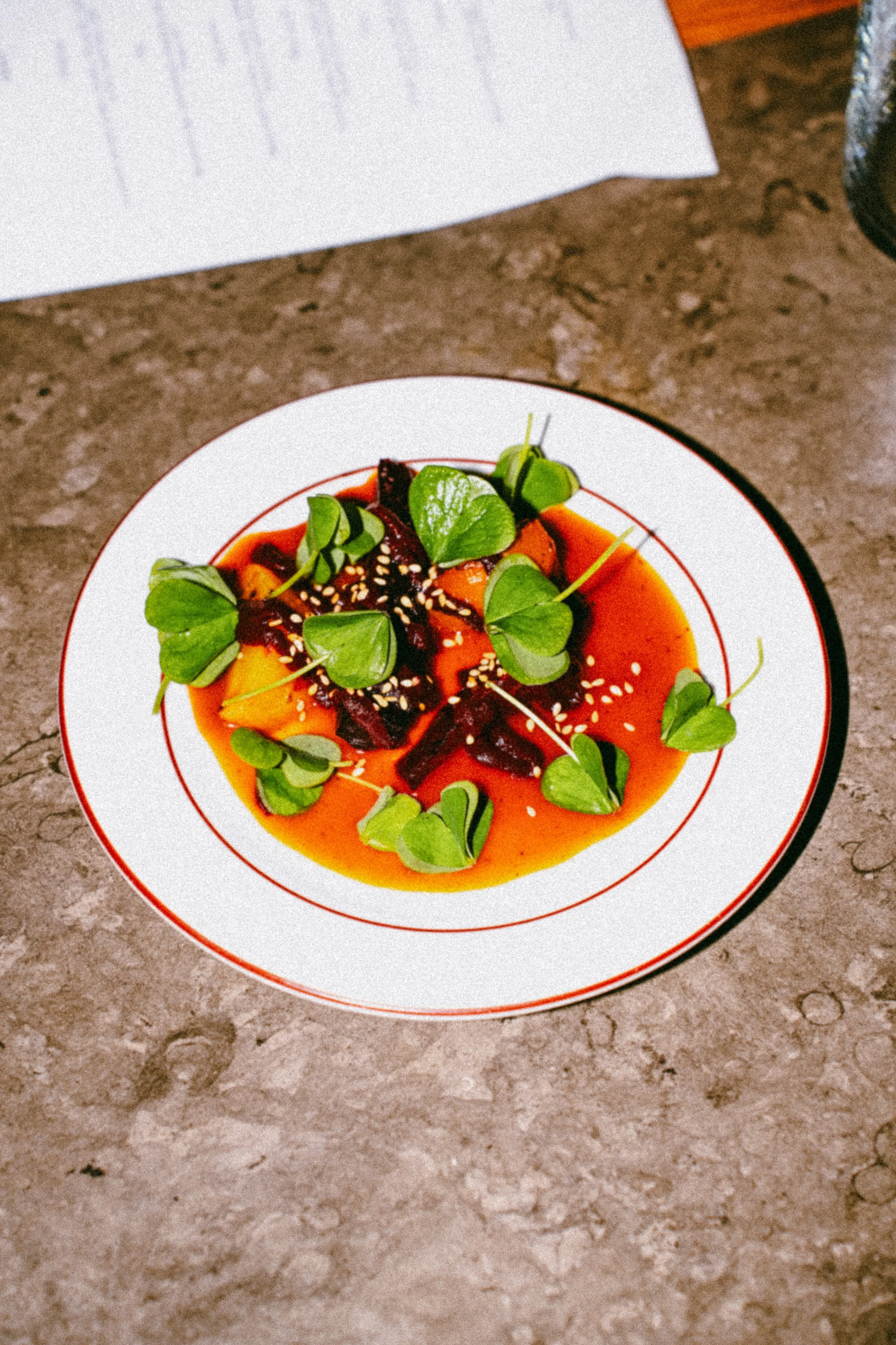 Plate of sliced vegetable, possibly beets and squash, topped with green microgreens and sprinkled with sesame seeds, served with a red sauce