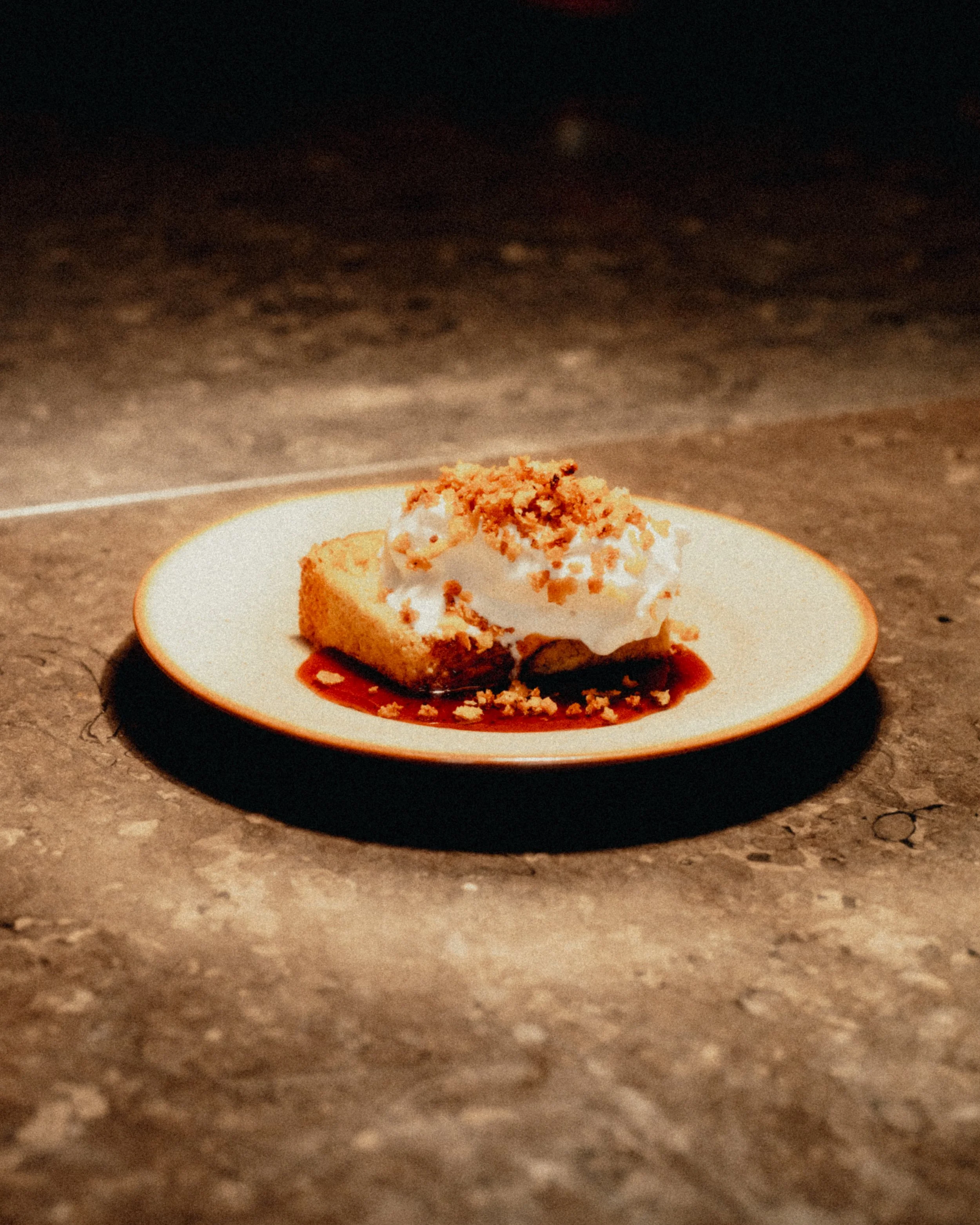 A dessert plate with a slice of cake topped with whipped cream and crushed nuts, served with a berry sauce on a textured dark surface.