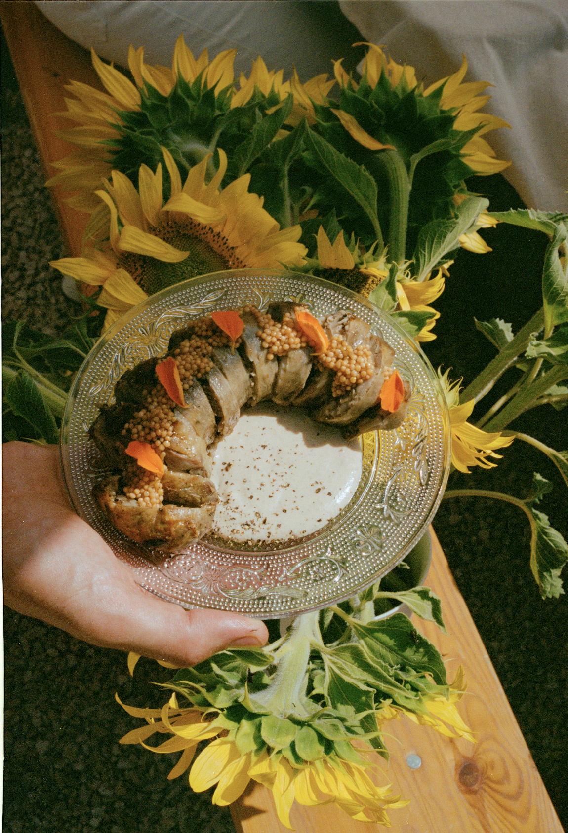 A decorative silver plate holds a baked stuffed chicken breast garnished with small orange flower petals and tiny round mustard seeds, held above a cluster of bright yellow sunflowers with green leaves.