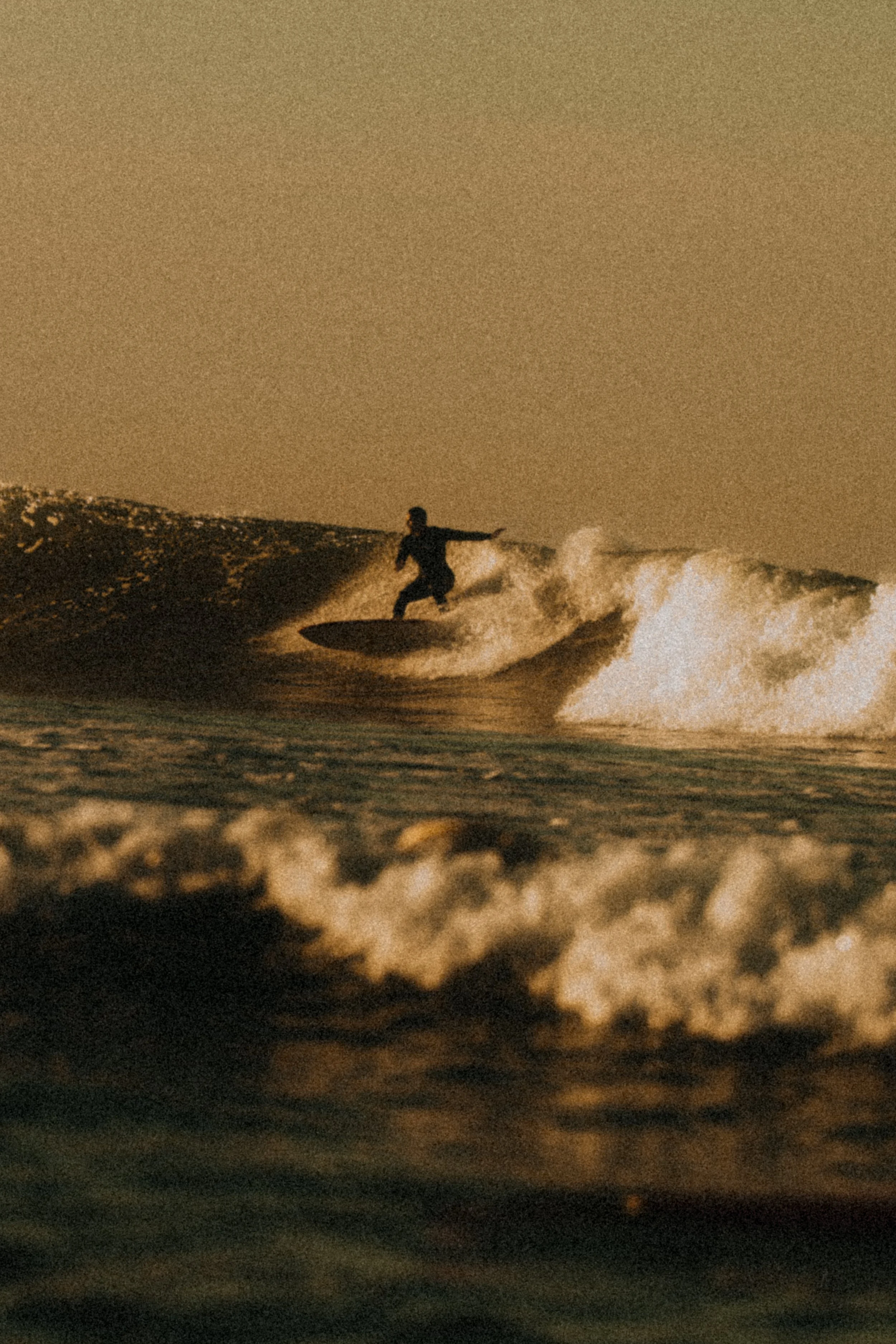A person surfing on a wave during sunset or sunrise, with a dark silhouette against the sky and water.