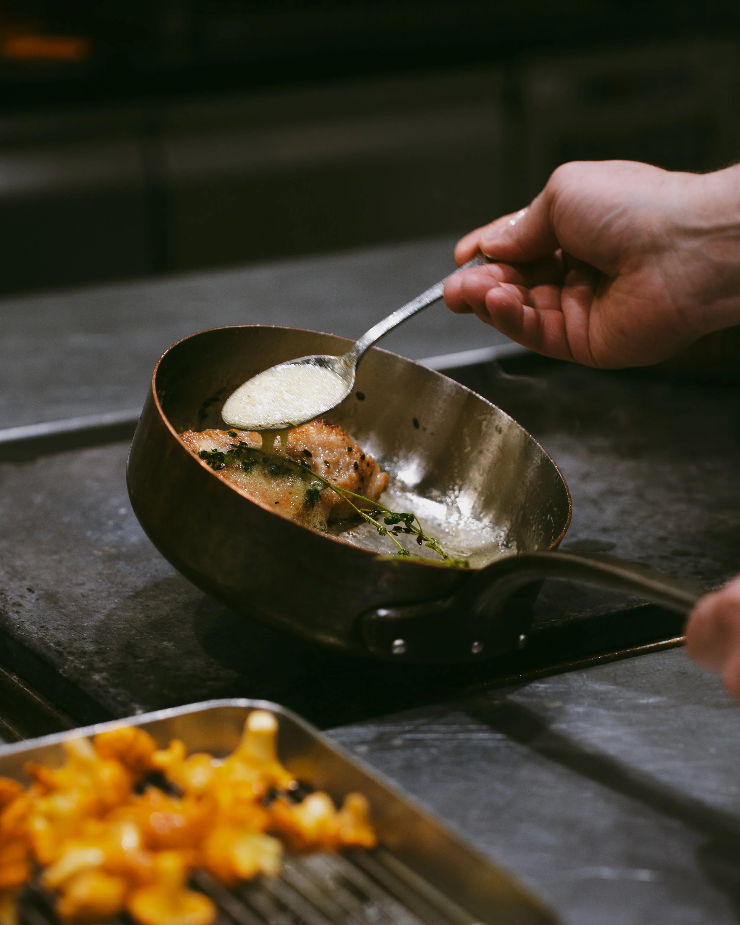 A chef adds a creamy sauce to a cooked piece of meat garnished with herbs in a small pan on a stovetop.