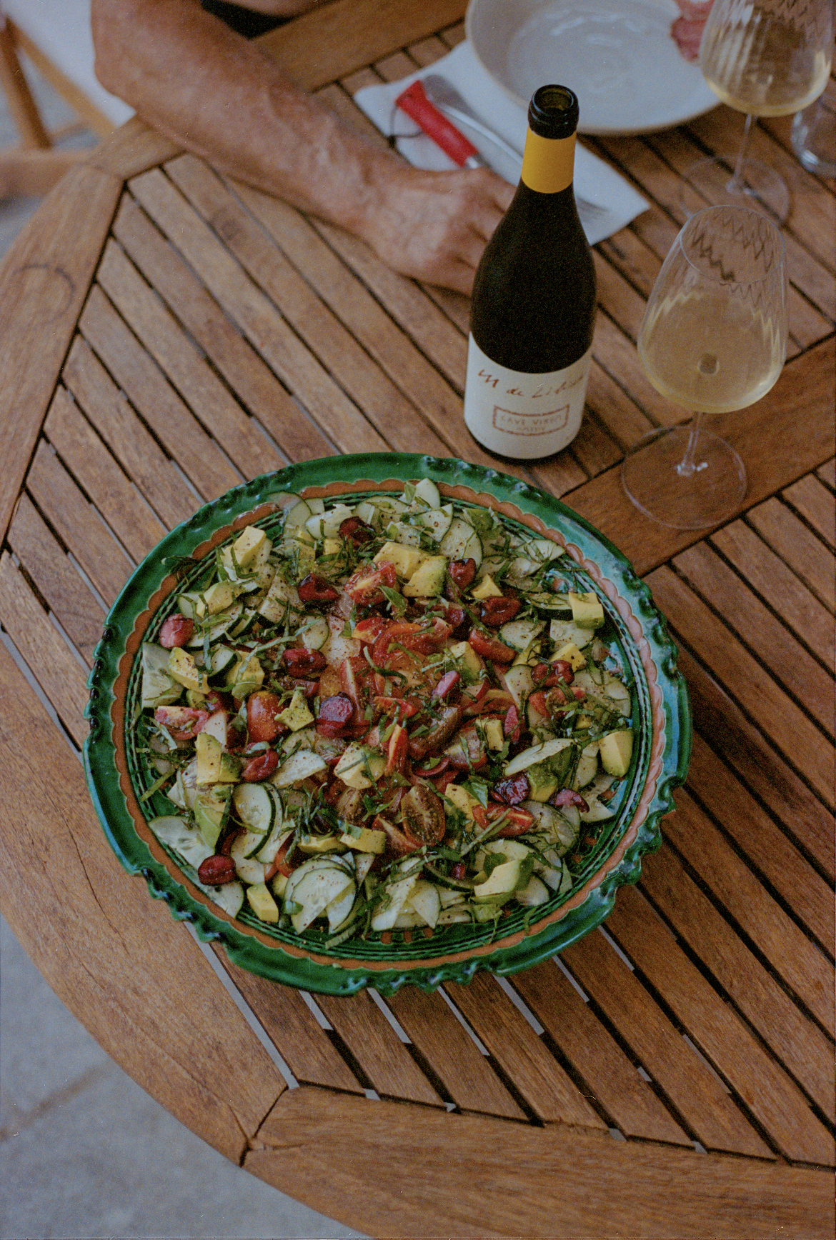Salad with chopped vegetables on a wooden table, with a bottle of wine, two wine glasses, and a person's arm in the background.