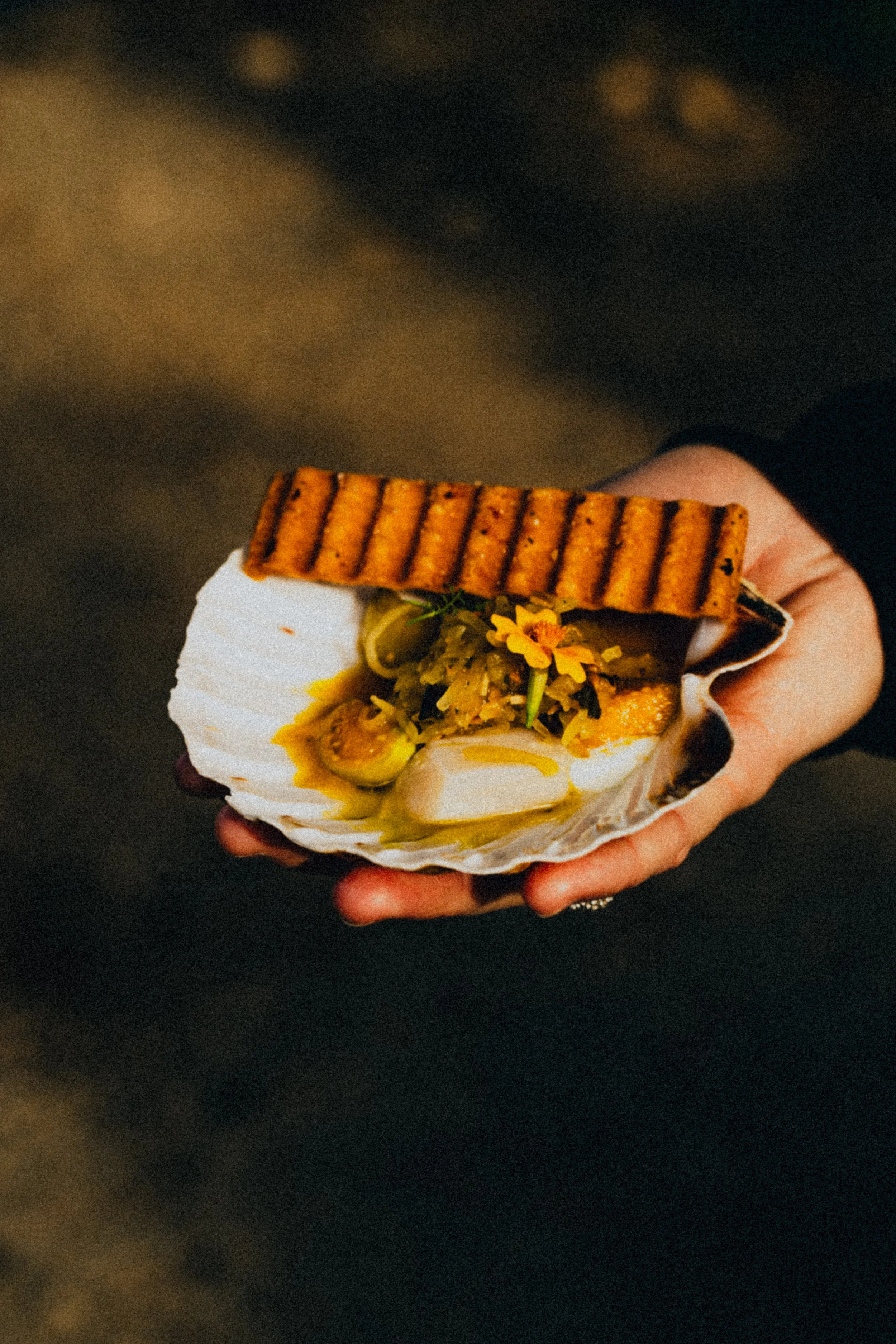 Person holding a scallop shell with grilled seafood, yellow sauce, and a grilled breadstick.