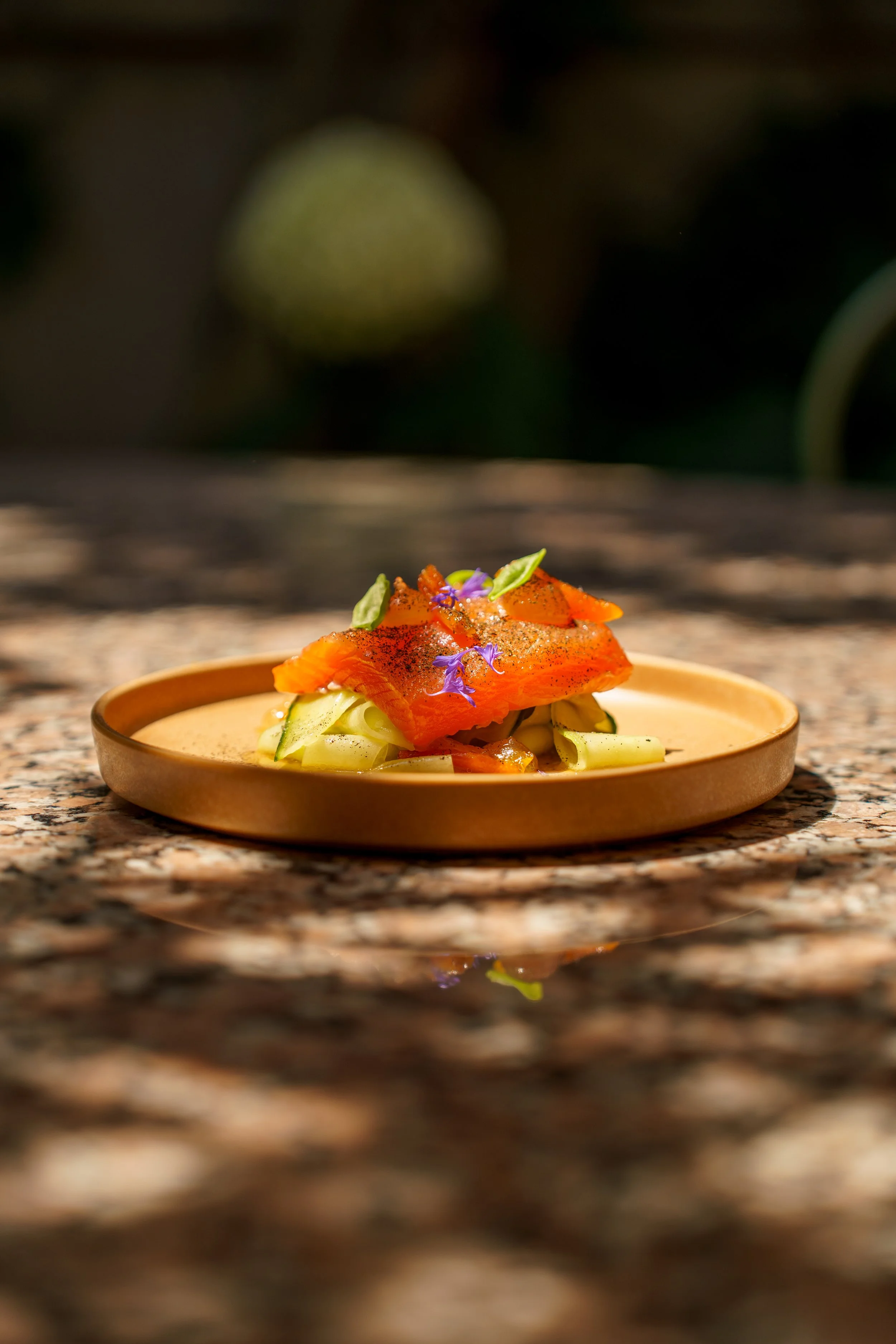 A plated dish with slices of smoked salmon, garnished with edible flowers and green herbs, on a round, shallow, tan-colored plate placed on a speckled granite countertop.