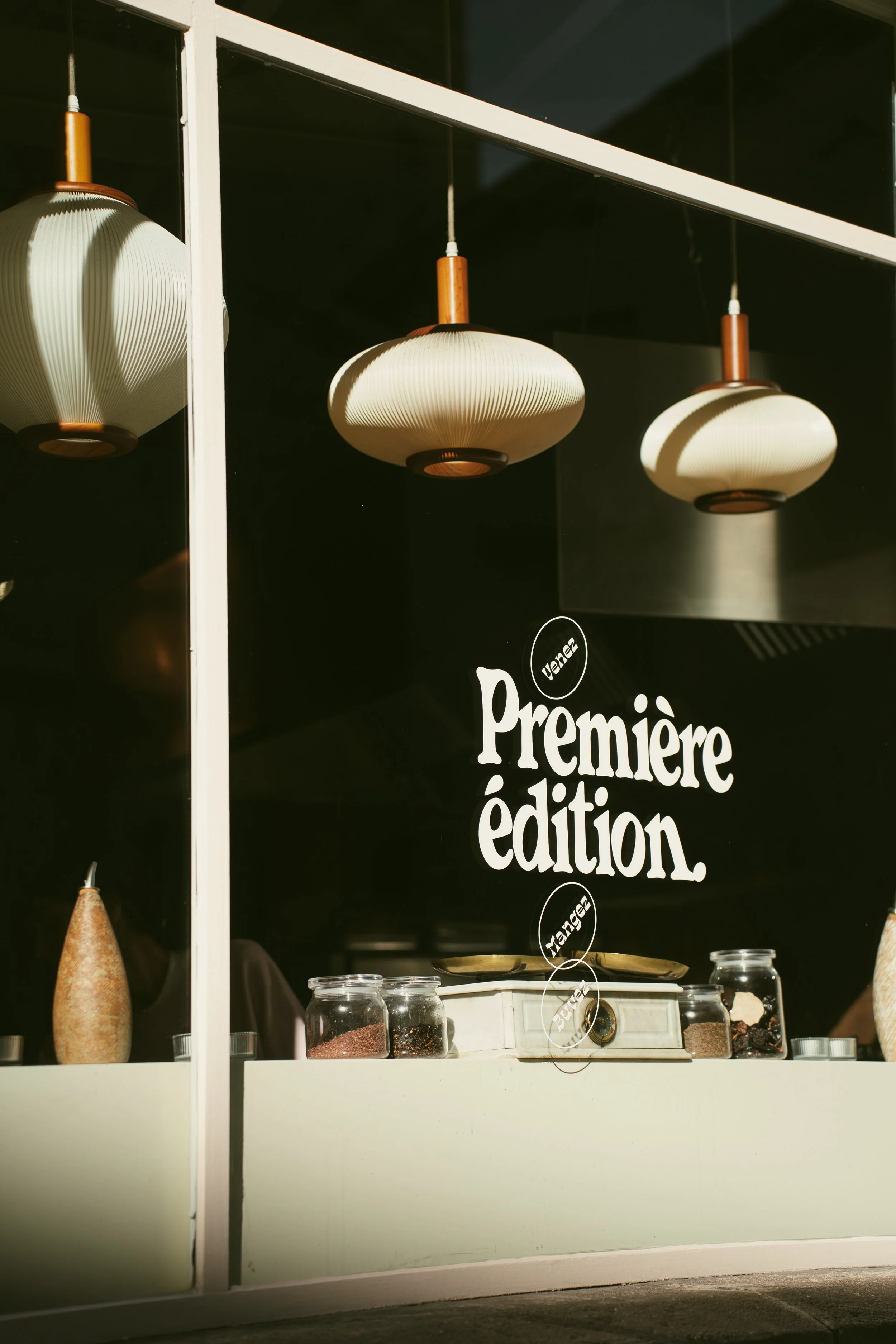 Interior of a shop window with three hanging pendant lights, jars of spices, and a vintage scale, with a sign reading "Première édition" in the background.