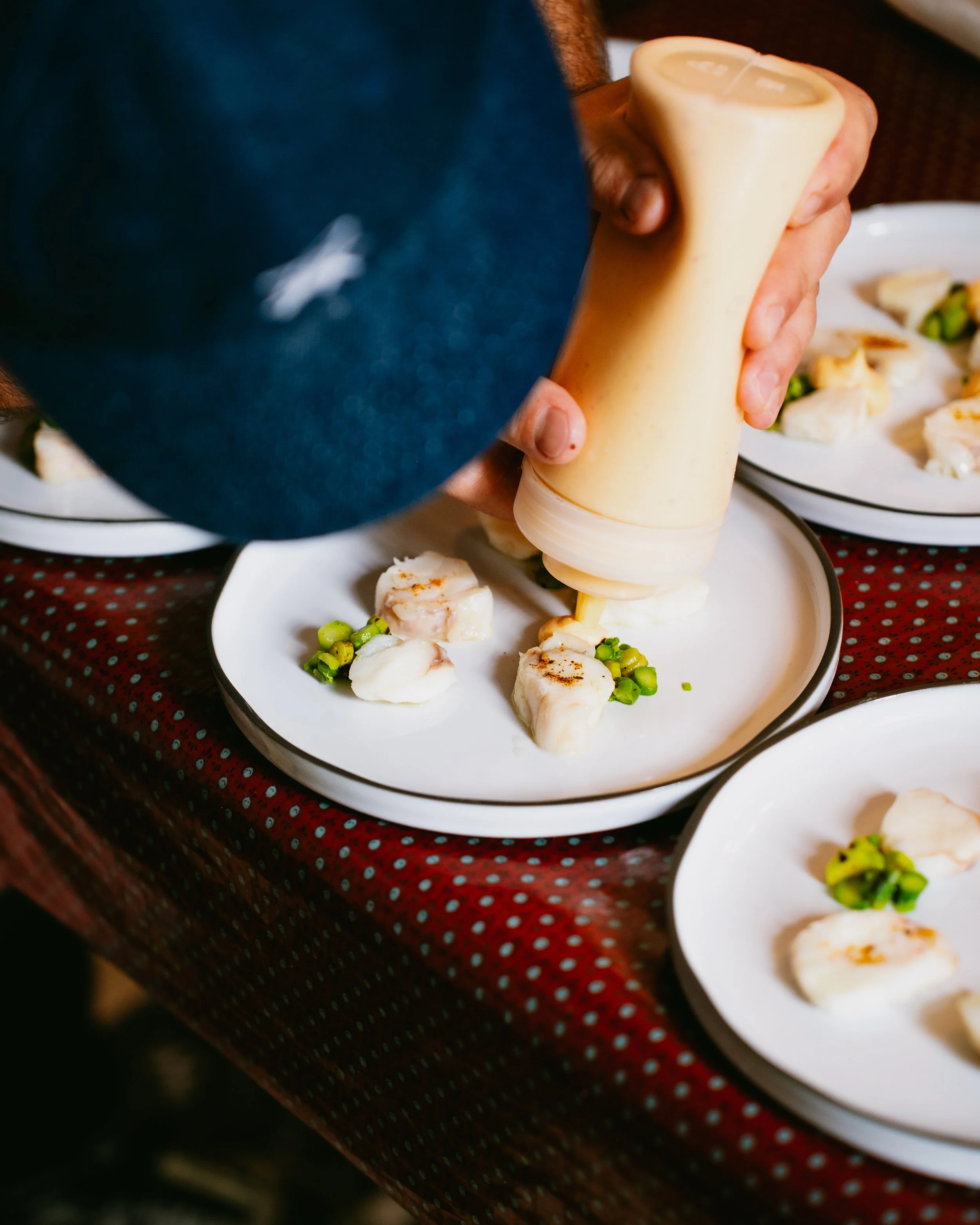 Person using a squeeze bottle to add sauce to a plate with cooked scallops and green peas, with plates of similar food in the background.