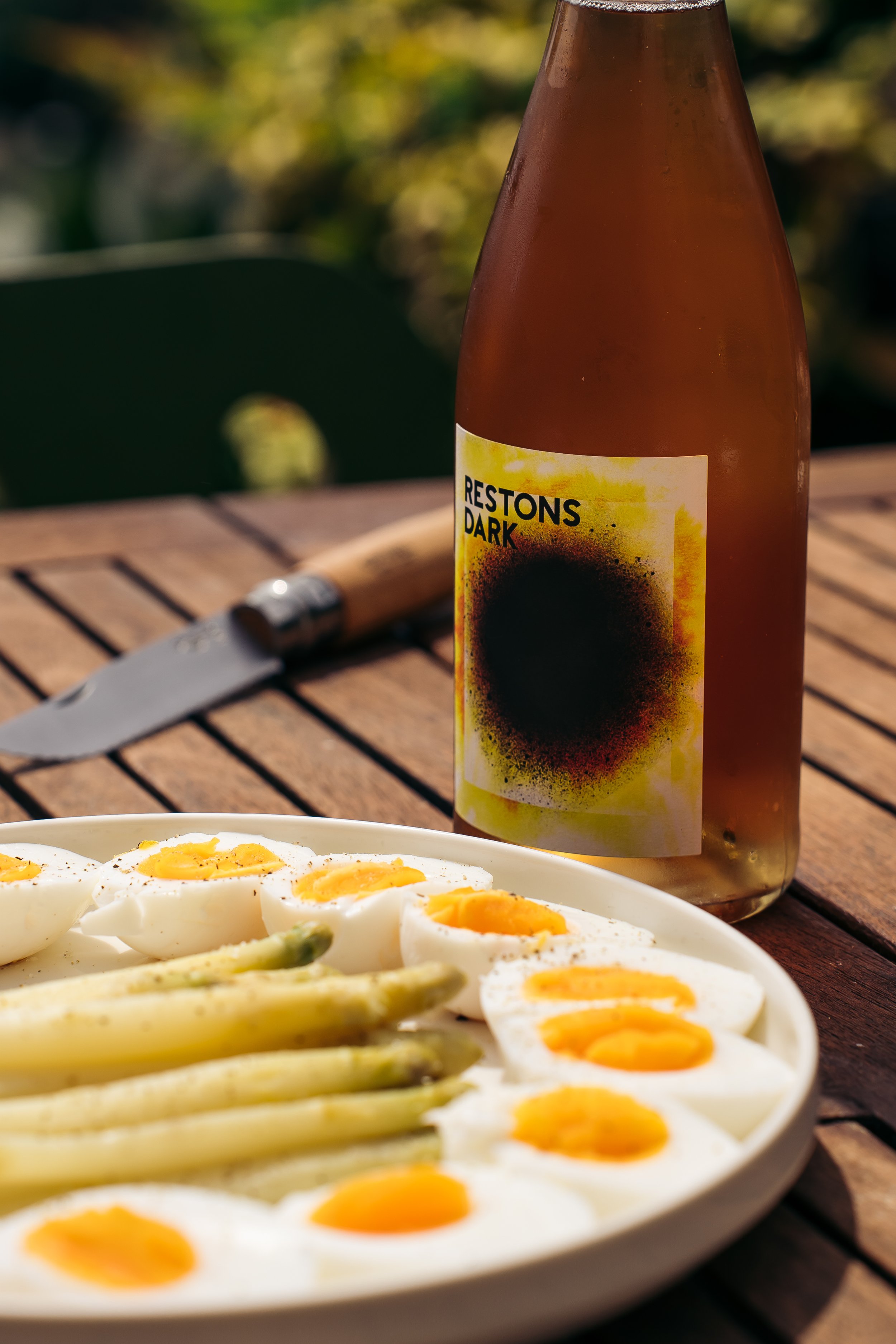A white plate with halved soft-boiled eggs and asparagus on a wooden table, with a bottle labeled 'Restons Dark' and a small knife in the background.