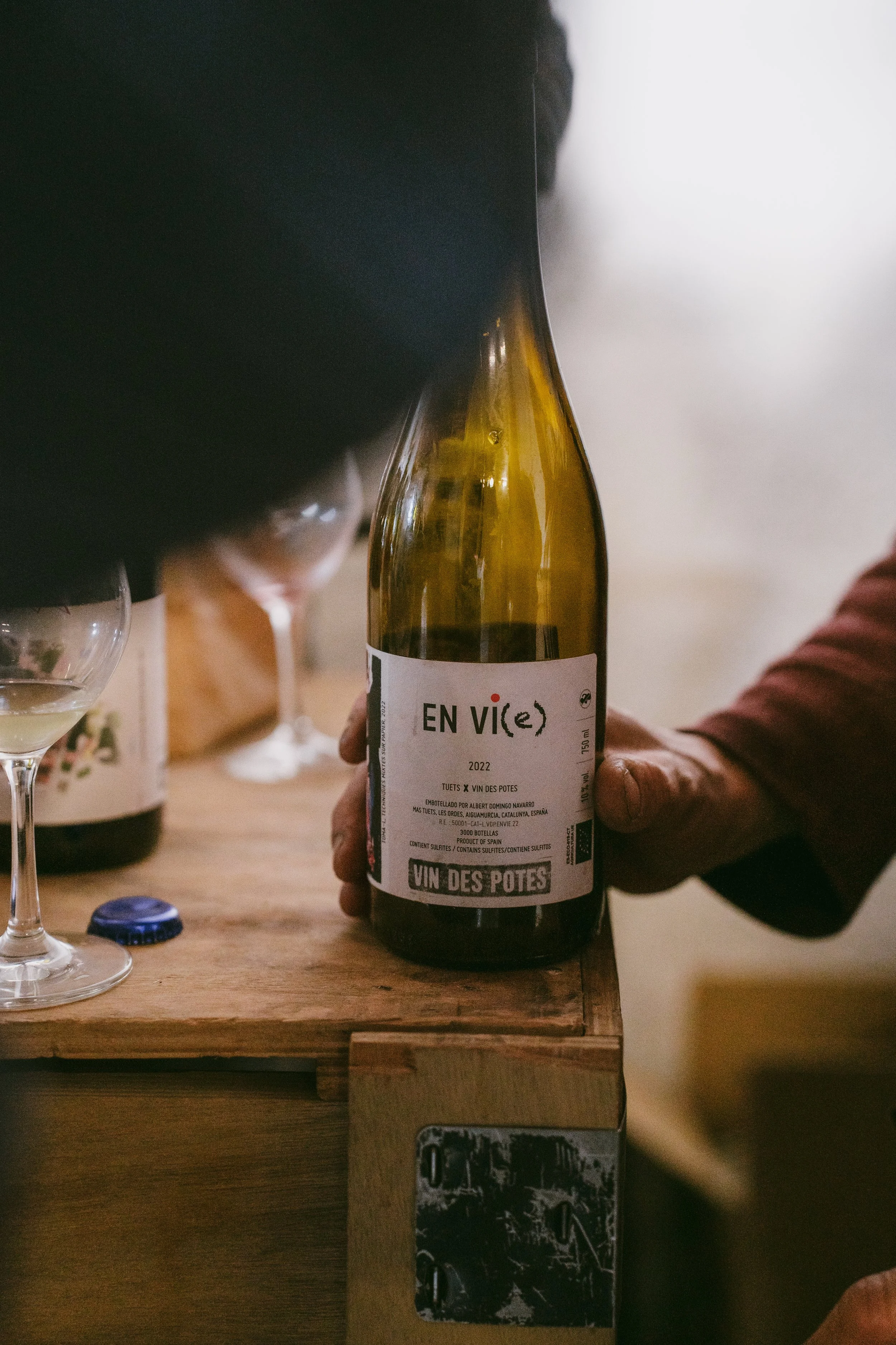 Person holding a bottle of wine labeled 'EN VIC(e) 2022' on a wooden table with glassware nearby.