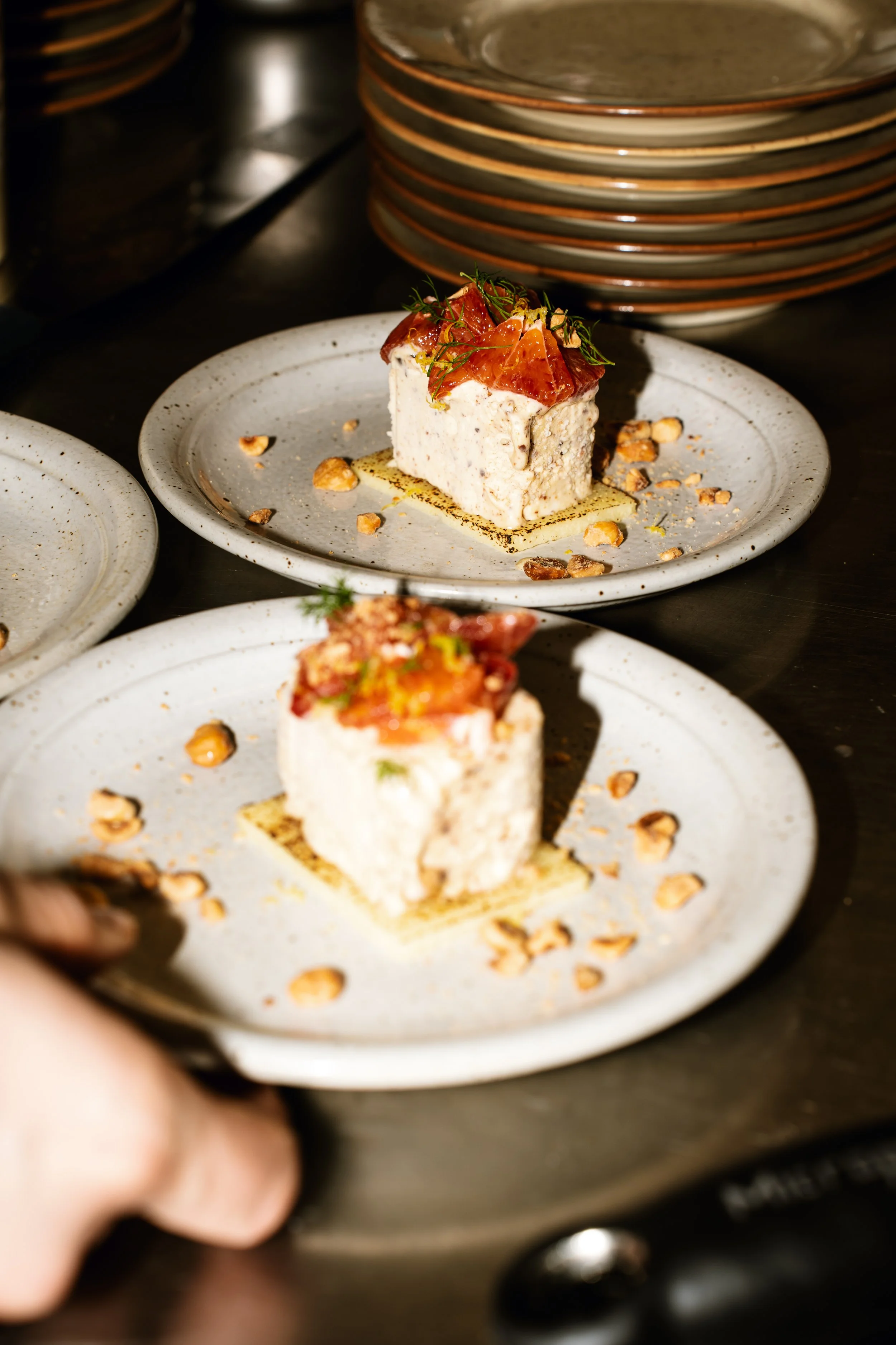 Two elegant dessert plates featuring small layered cakes topped with thin slices of cured meat, herbs, and garnished with chopped nuts.