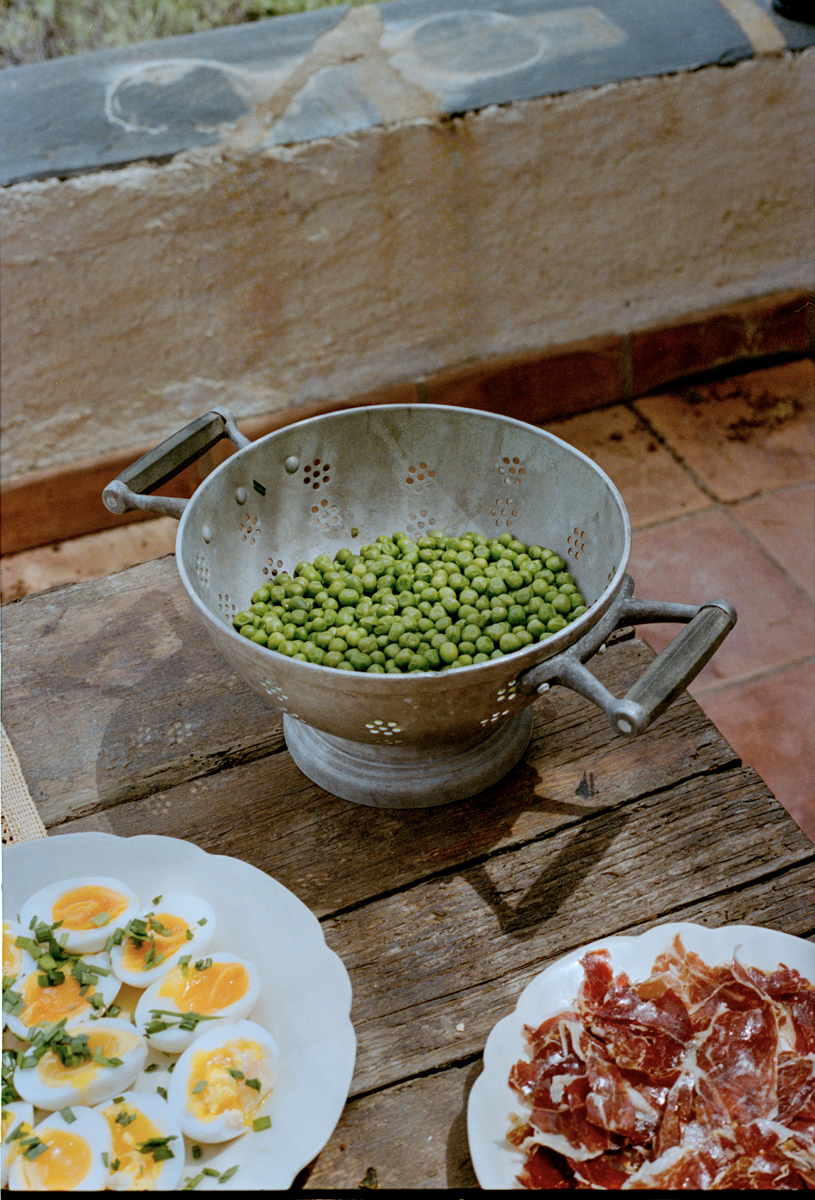 A metal colander filled with green peas on a rustic wooden table, with plates of halved boiled eggs garnished with chopped herbs and a pile of sliced cured ham nearby.