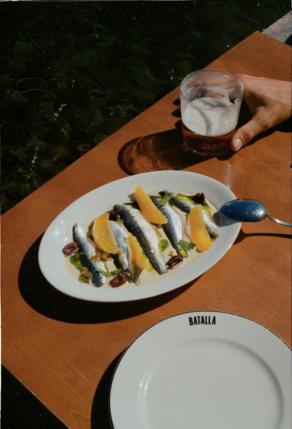 A plate of sardines with orange slices and herbs on a wooden table next to a glass of beer and an empty plate with 'BATALLA' written on it.