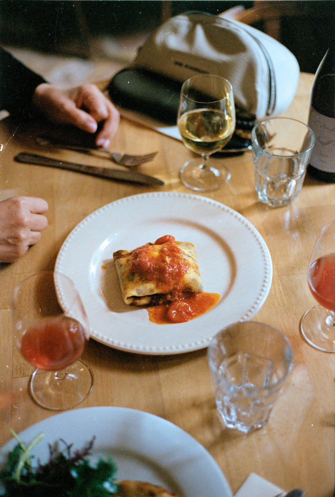 A plate of baked pasta with tomato sauce on a wooden dining table, surrounded by glasses of white and rosé wine, with a person's hand in the background.