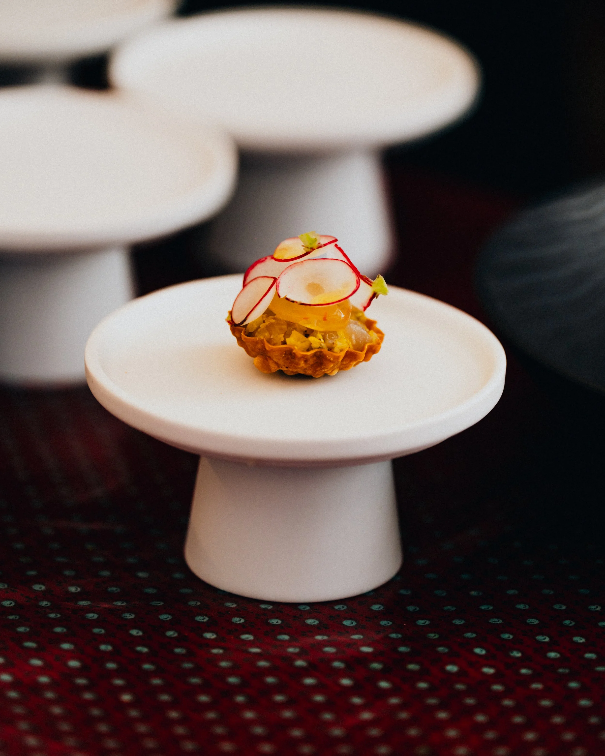 A miniature tart topped with thinly sliced radishes, a yellow topping, and garnished with tiny green herbs, placed on a white ceramic stand.